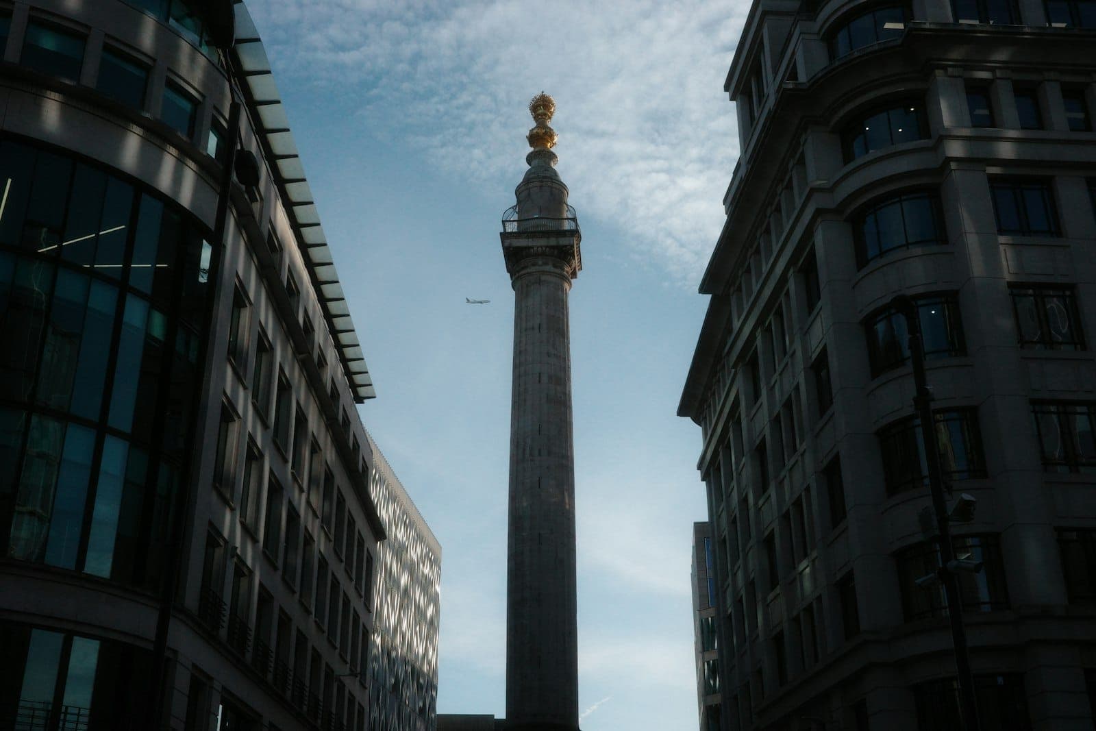 Monument to the Great Fire of London wznosi się między dwoma nowoczesnymi budynkami biurowymi pod jasnym błękitnym niebem z rozproszonymi chmurami.