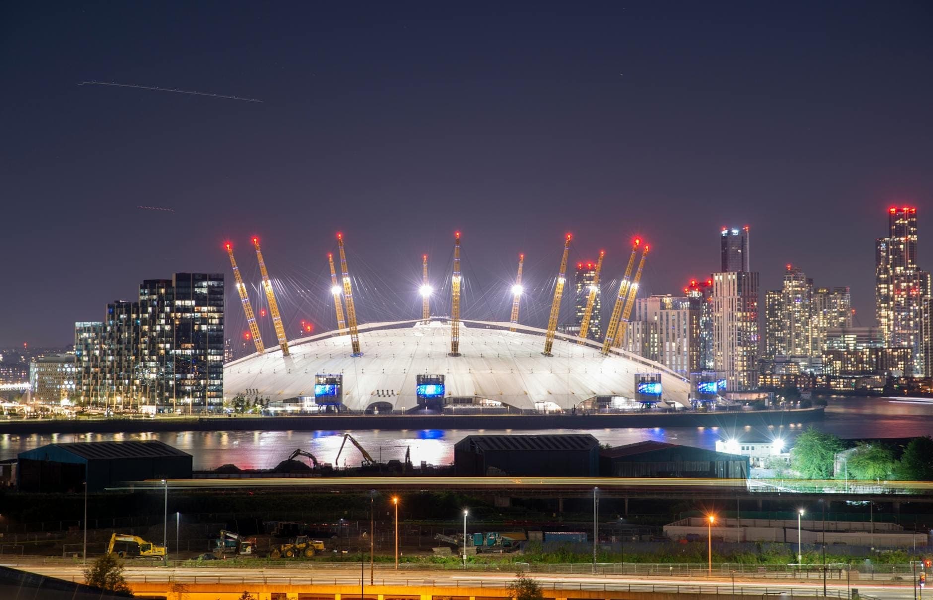 The O2 Arena oświetlona nocą z londyńskimi wieżowcami w tle, widok znad rzeki pod czystym niebem.