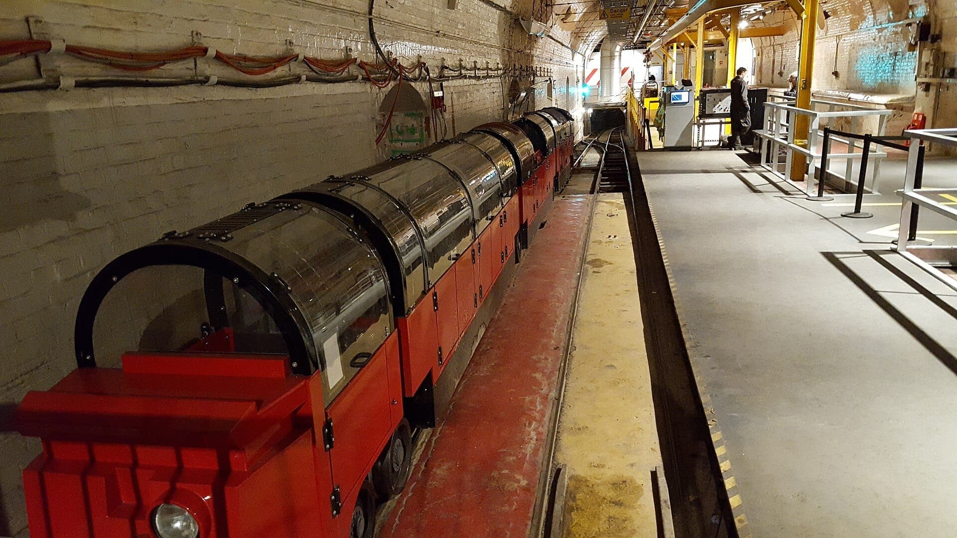 Kereta merah mini Mail Rail di The Postal Museum, London, terparkir di dalam stasiun terowongan bawah tanah dengan pengunjung di peron.