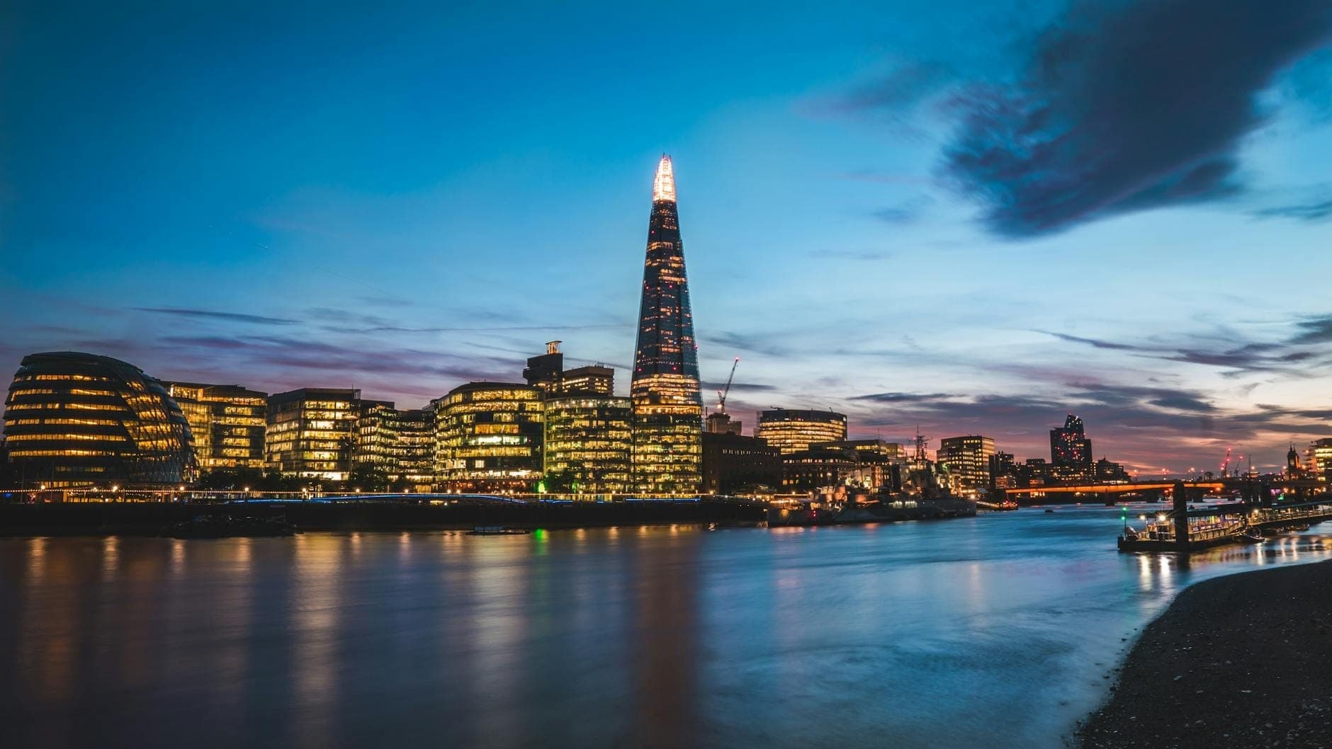 Le gratte-ciel The Shard illuminé au crépuscule, dominant la skyline de Londres avec ses lumières se reflétant dans la Tamise, sous un ciel vibrant aux teintes bleues et violettes.