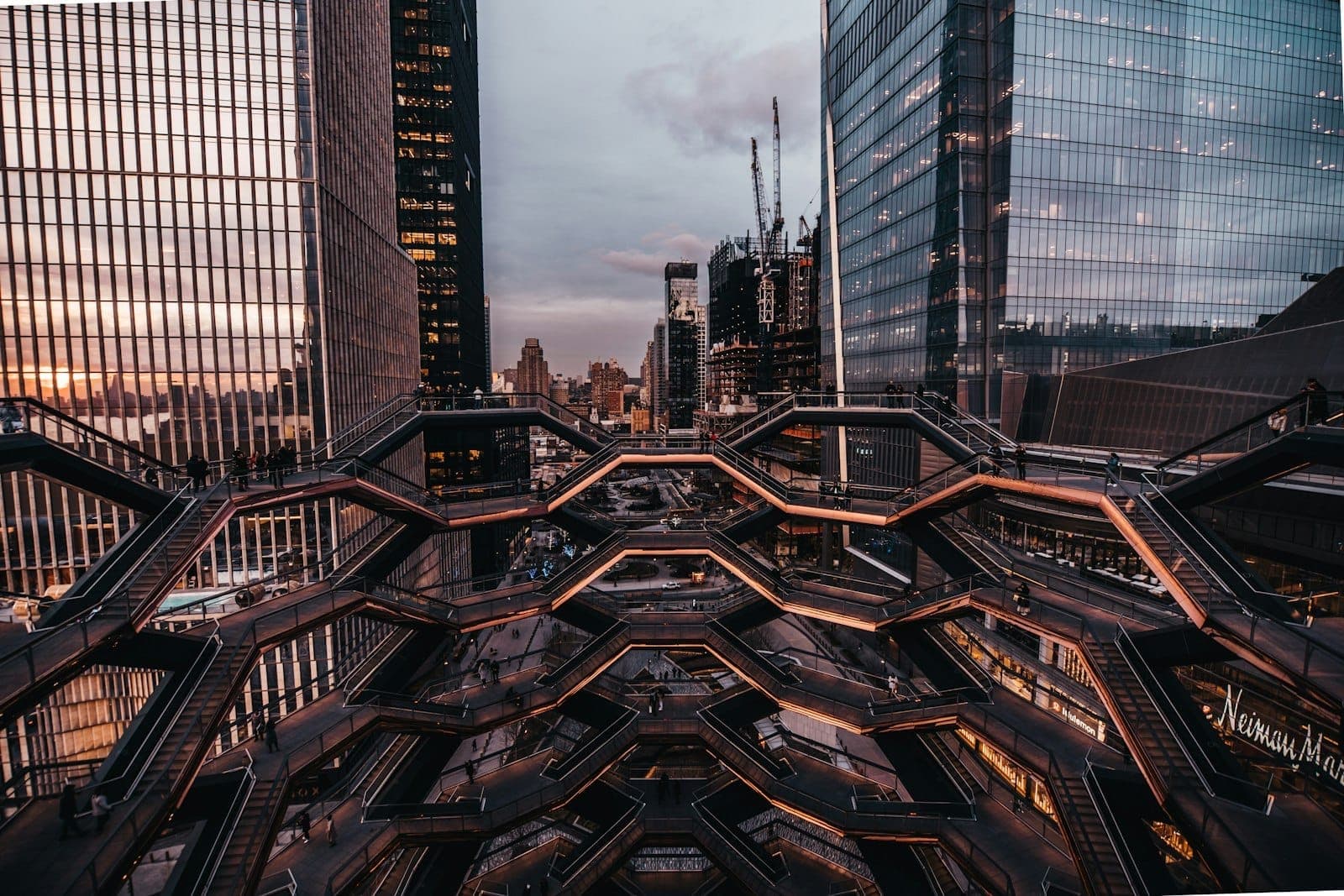 บันไดทองแดงรูปทรงเรขาคณิตของ The Vessel กับตึกสูง Hudson Yards ยามเย็น เห็นวิวสุดตระการตาของโครงสร้างและเมืองรอบข้าง