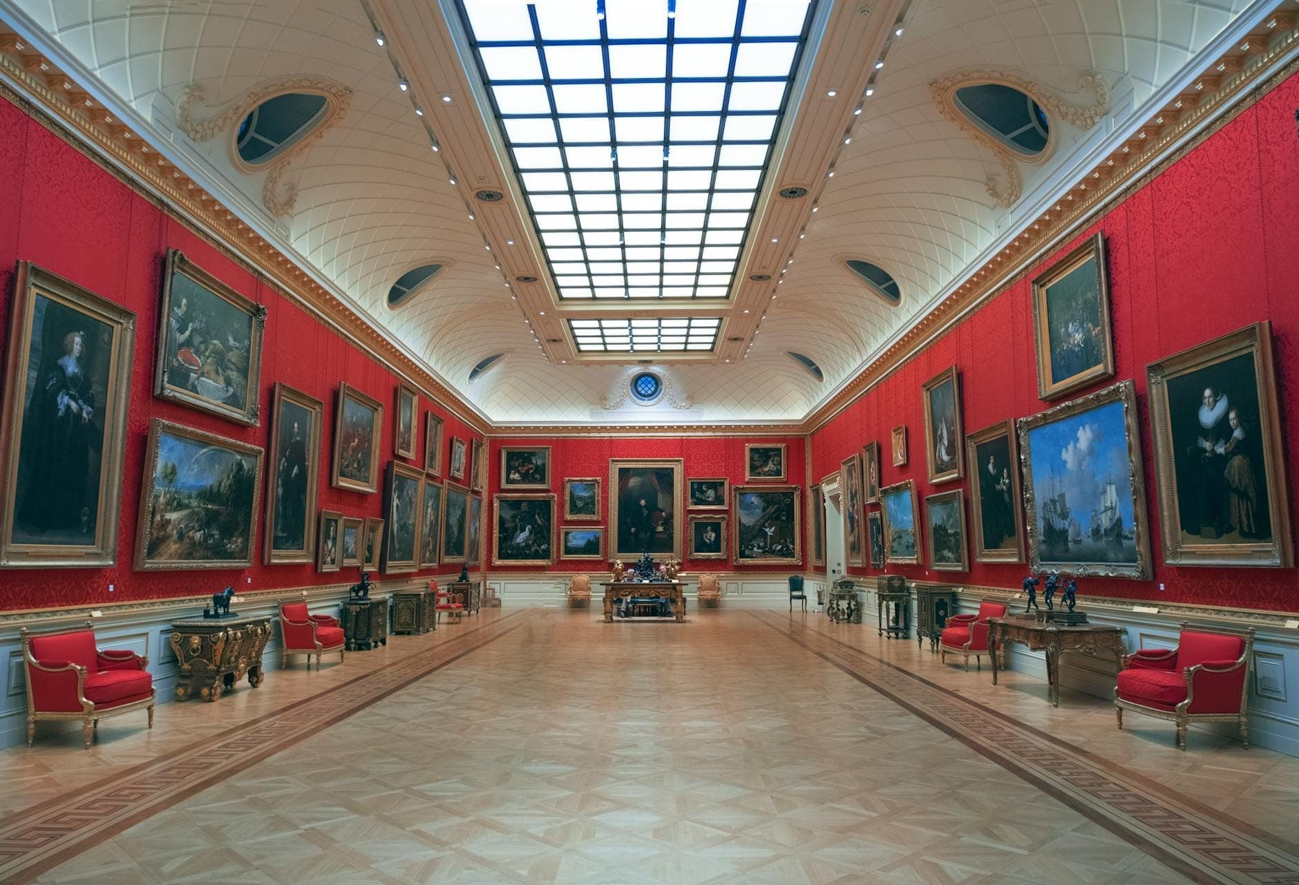 A grand, lavish gallery hall inside the Wallace Collection in London, featuring red walls lined with Old Master paintings, ornate furniture, and a bright glass ceiling.