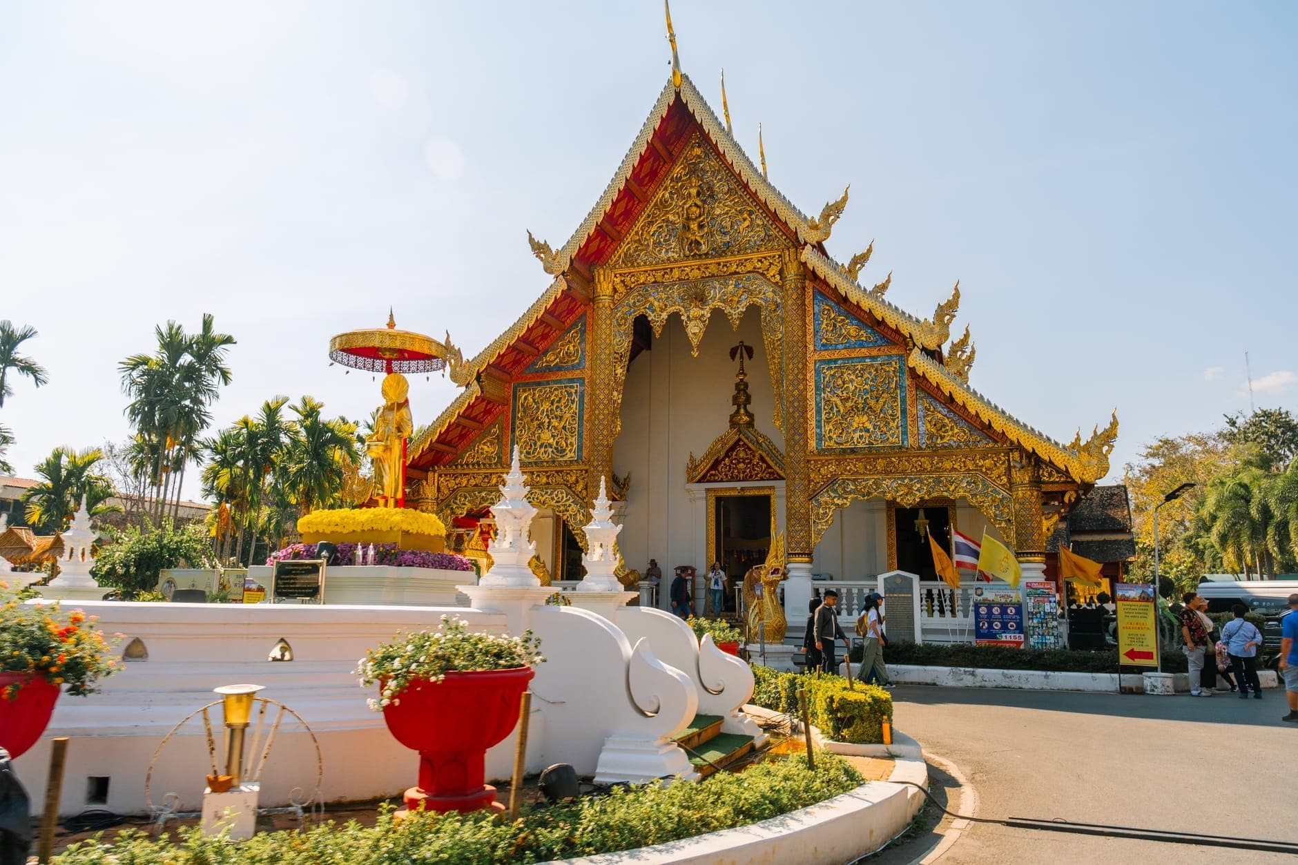 Zdobna złota świątynia w Chiang Mai w Tajlandii, z ludźmi spacerującymi wokół wejścia i bujną zielenią otaczającą budynek w słoneczny dzień.