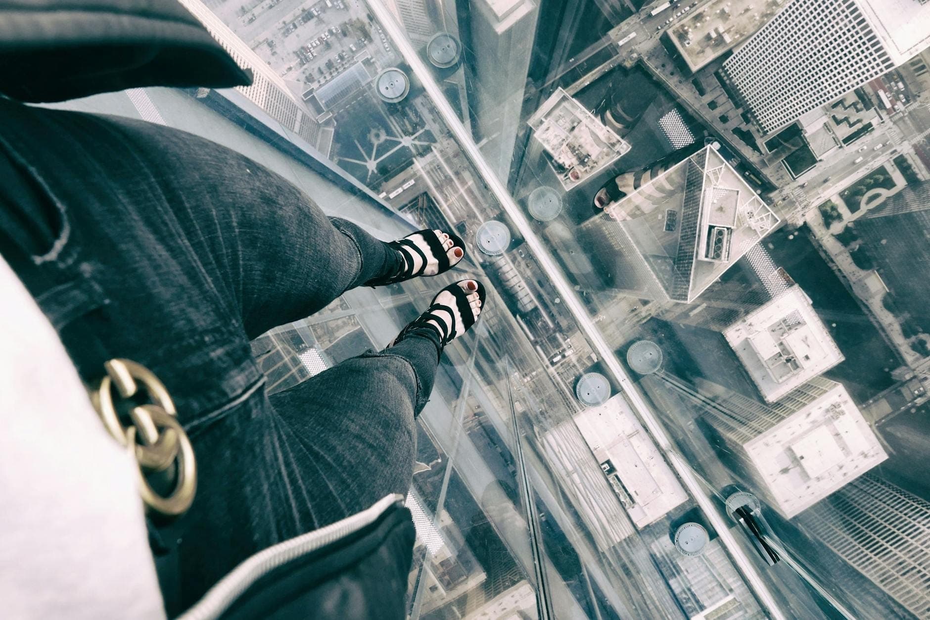 Person standing on glass observation ledge high above Chicago skyline, showing view down to streets and rooftops below.