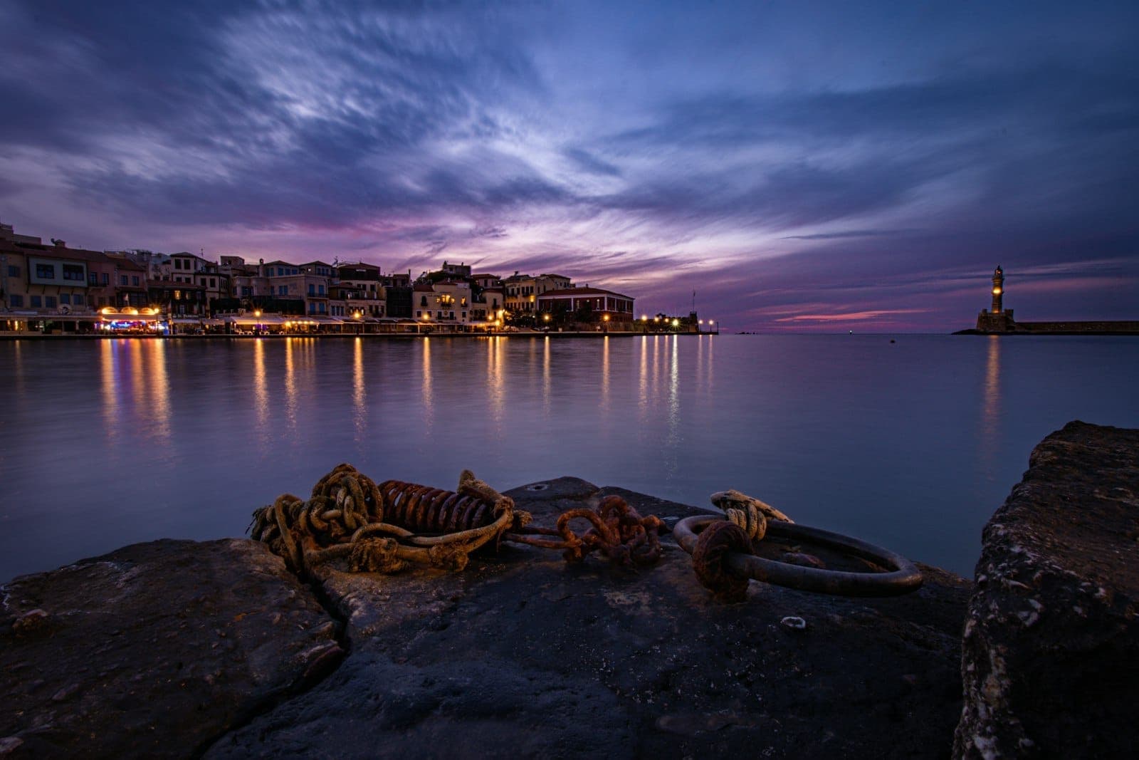 Puerto veneciano de Chania al atardecer con un destacado faro, edificios históricos frente al mar y dramáticos reflejos del cielo nocturno en el agua.
