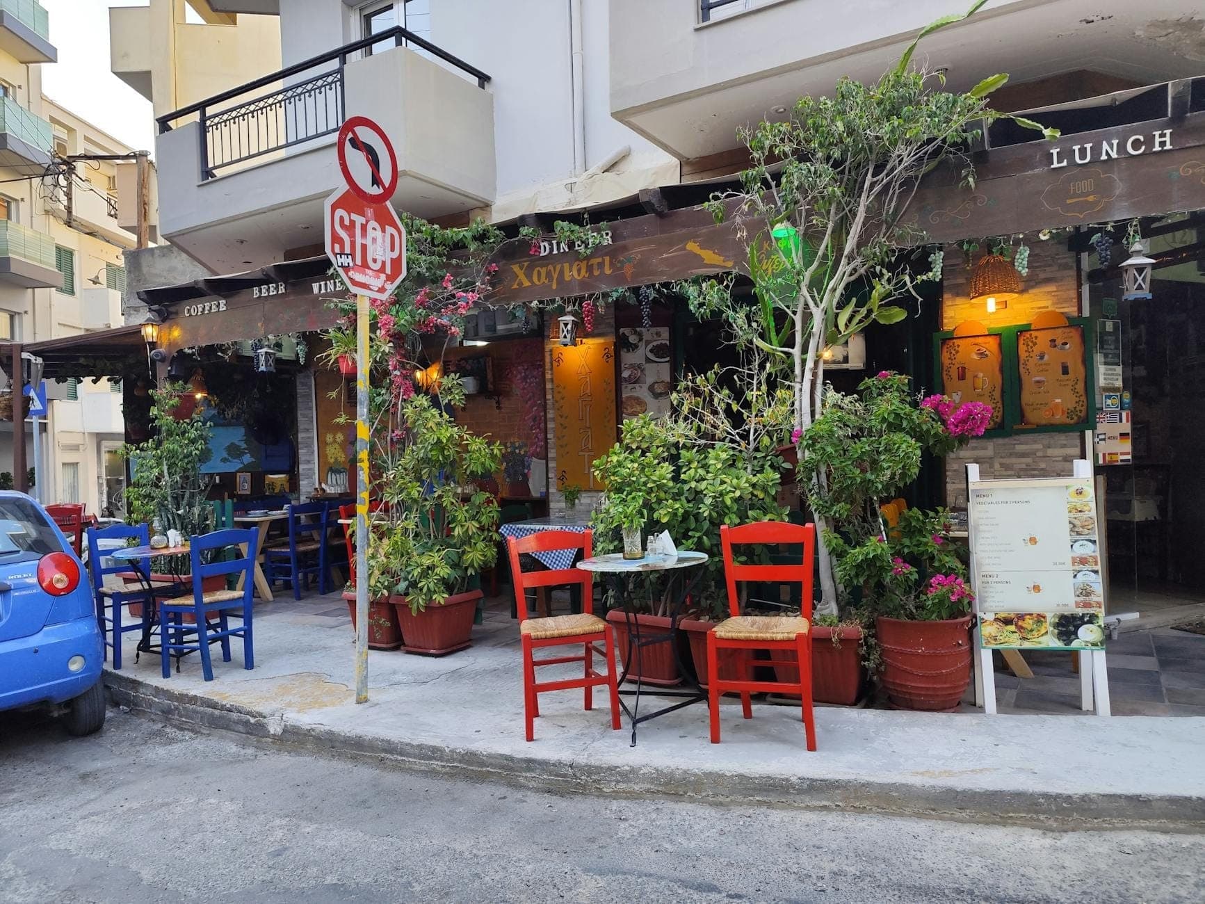 Mesas y sillas coloridas al aire libre en una acogedora taberna cretense con exuberantes plantas, creando un ambiente acogedor de cultura gastronómica local.