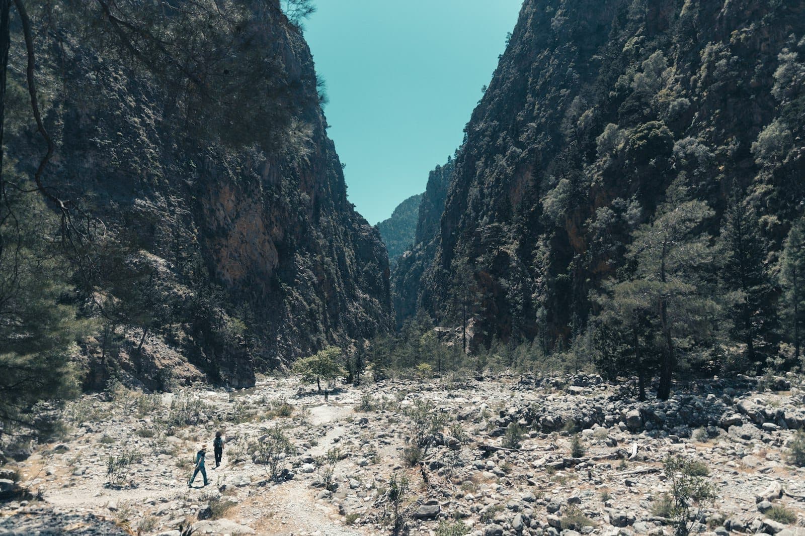 Dos senderistas caminan por un desfiladero rocoso con escarpadas paredes de acantilado y árboles dispersos bajo un cielo azul despejado en Creta.