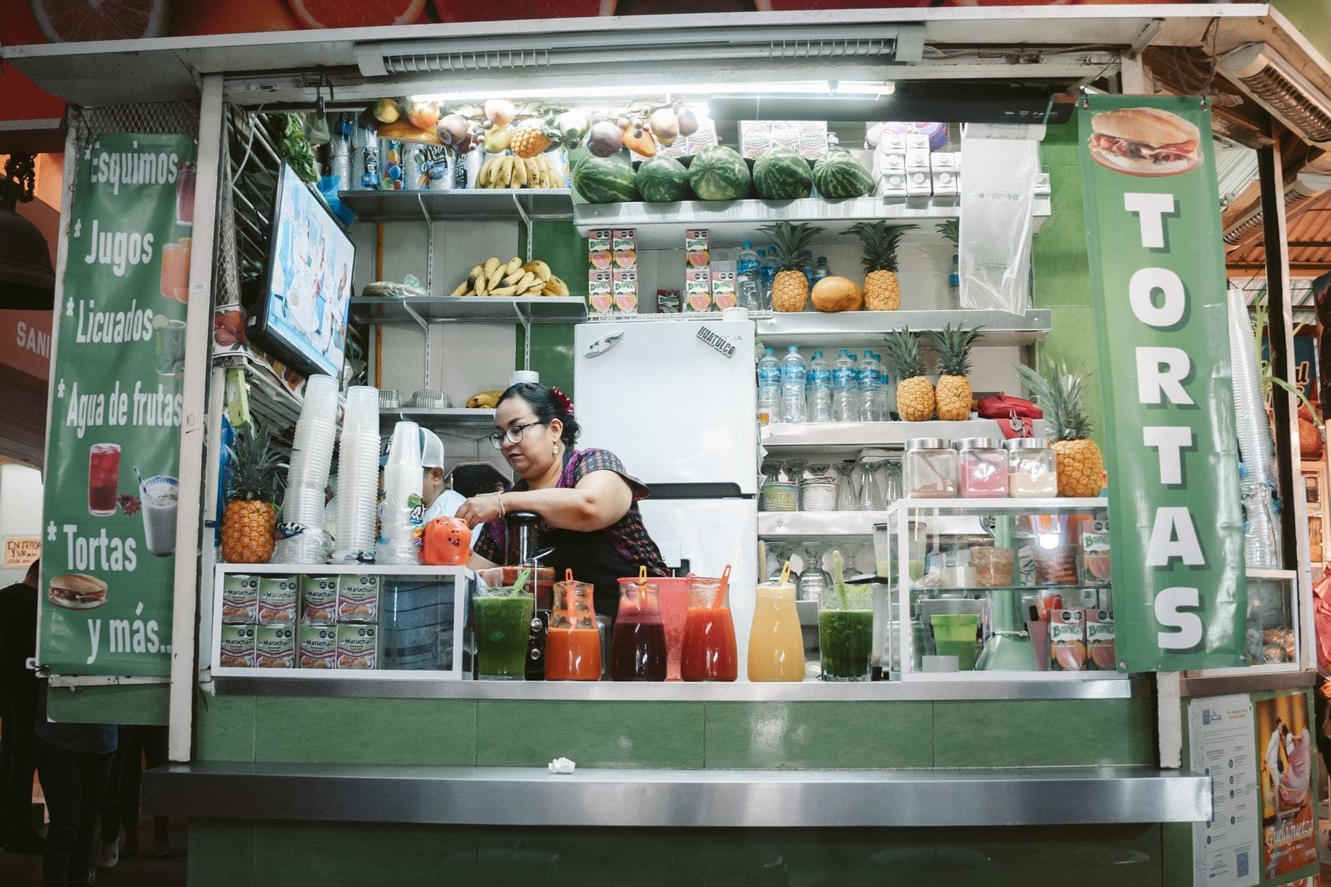 แผงขายอาหารในตลาดของกวาดาลาฮาราที่จำหน่าย tortas น้ำผลไม้ และผลไม้สด โดยมีผู้หญิงกำลังเตรียมอาหารอยู่หลังเคาน์เตอร์