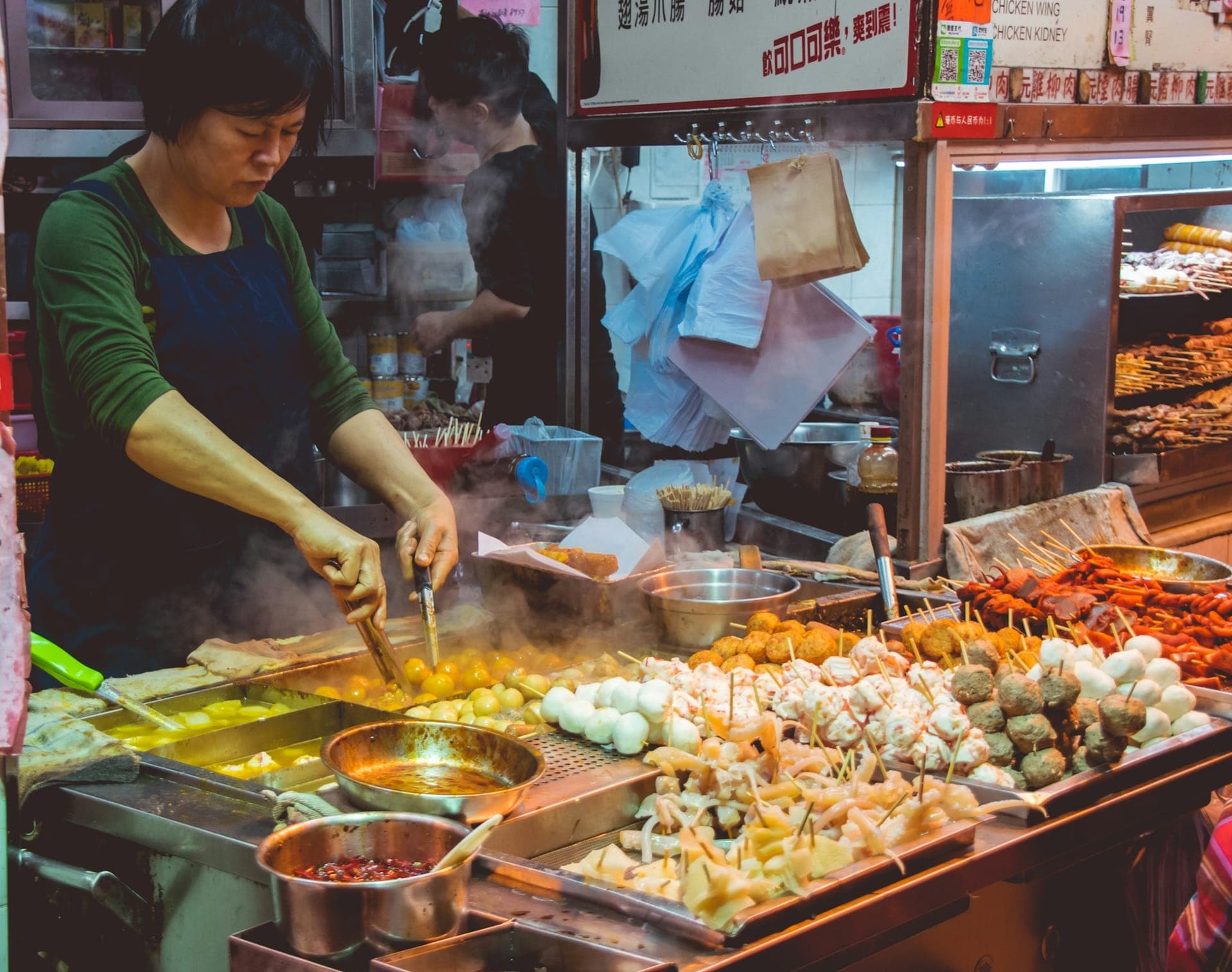 Hong Kong street food stall at a night market, vendor grilling fish balls, meat skewers, and offal over steaming woks behind a crowded counter filled with bamboo skewers and trays of ingredients.