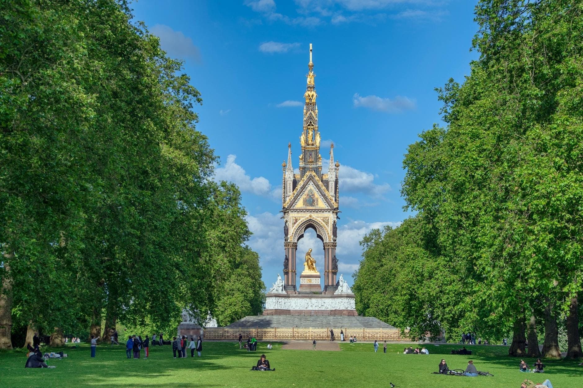 Personas descansando y paseando sobre el césped frente al ornamentado Albert Memorial en los Kensington Gardens de Londres, rodeado de frondosos árboles verdes.