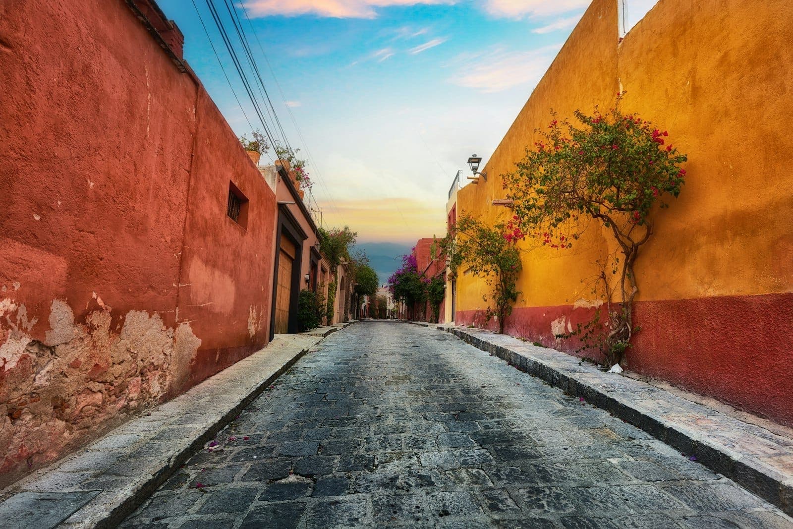 Calle adoquinada flanqueada de vibrantes paredes rojas y amarillas, con un árbol y un suave cielo al atardecer.
