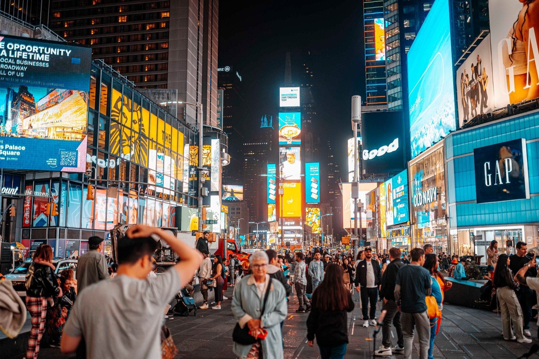 ฝูงชนที่ Times Square ในยามค่ำคืนพร้อมป้ายโฆษณาและแสงไฟสว่างจ้า สะท้อนถึงบรรยากาศอันคึกคักของชีวิตกลางคืนในนครนิวยอร์ก