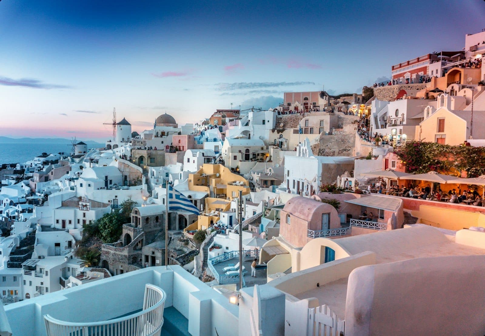 Santorins ikonische weiß getünchte Häuser mit blauen Kuppeln auf Klippen im Sonnenuntergang, mit Blick auf das Meer und pastellfarbenem Himmel.