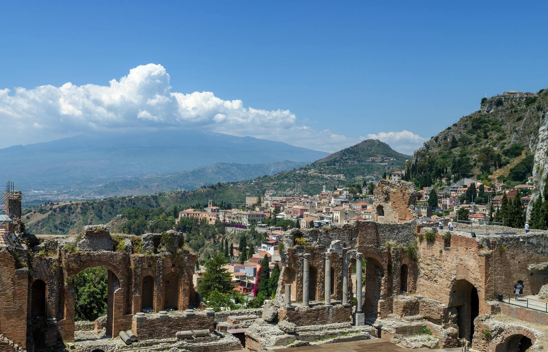 Reruntuhan teater Yunani kuno di Taormina dengan Gunung Etna dan garis pantai Sisilia di latar belakang pada hari yang cerah.