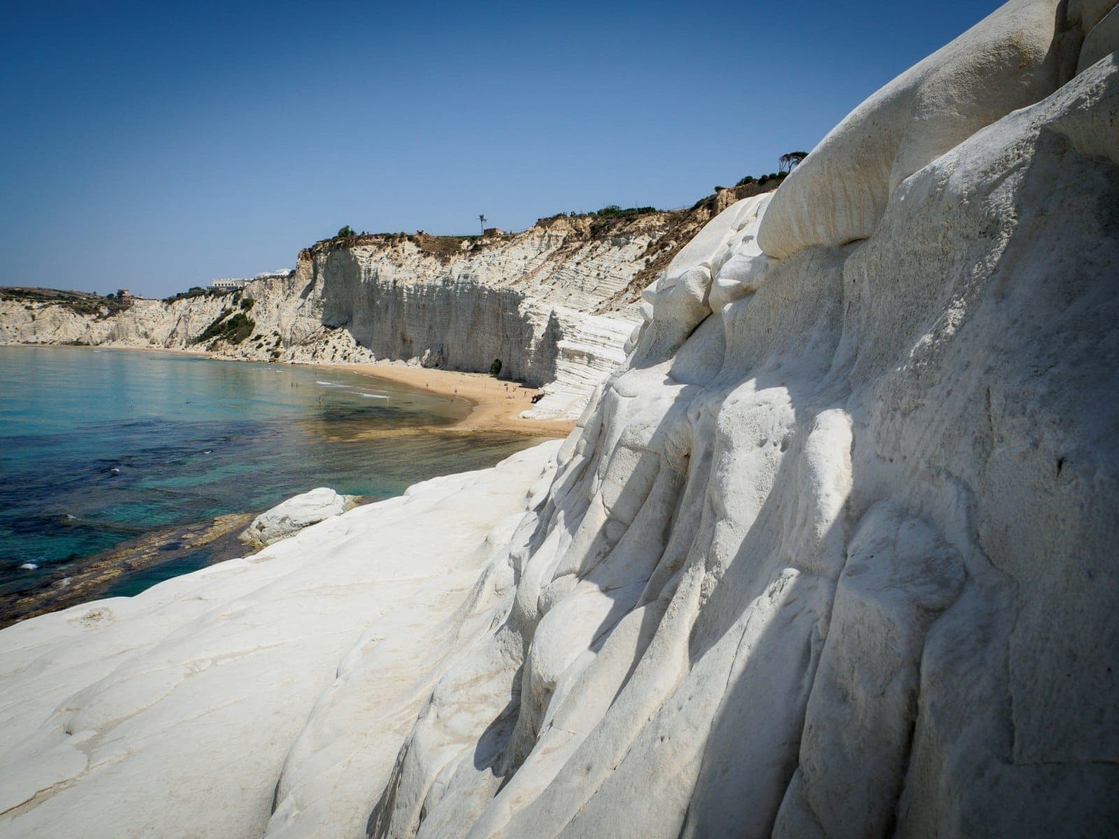 Tebing marly putih Scala dei Turchi dengan air biru jernih dan pantai berpasir di bawahnya di pesisir Sisilia.