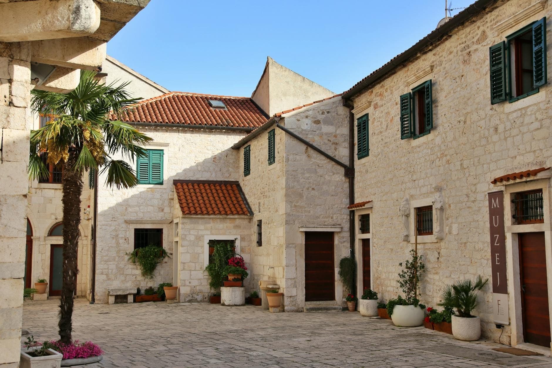 Cour ensoleillée en pierre avec des plantes en pots et des bâtiments, dont un panneau visible indiquant 'MUZEJ' désignant un musée à Split, Croatie.