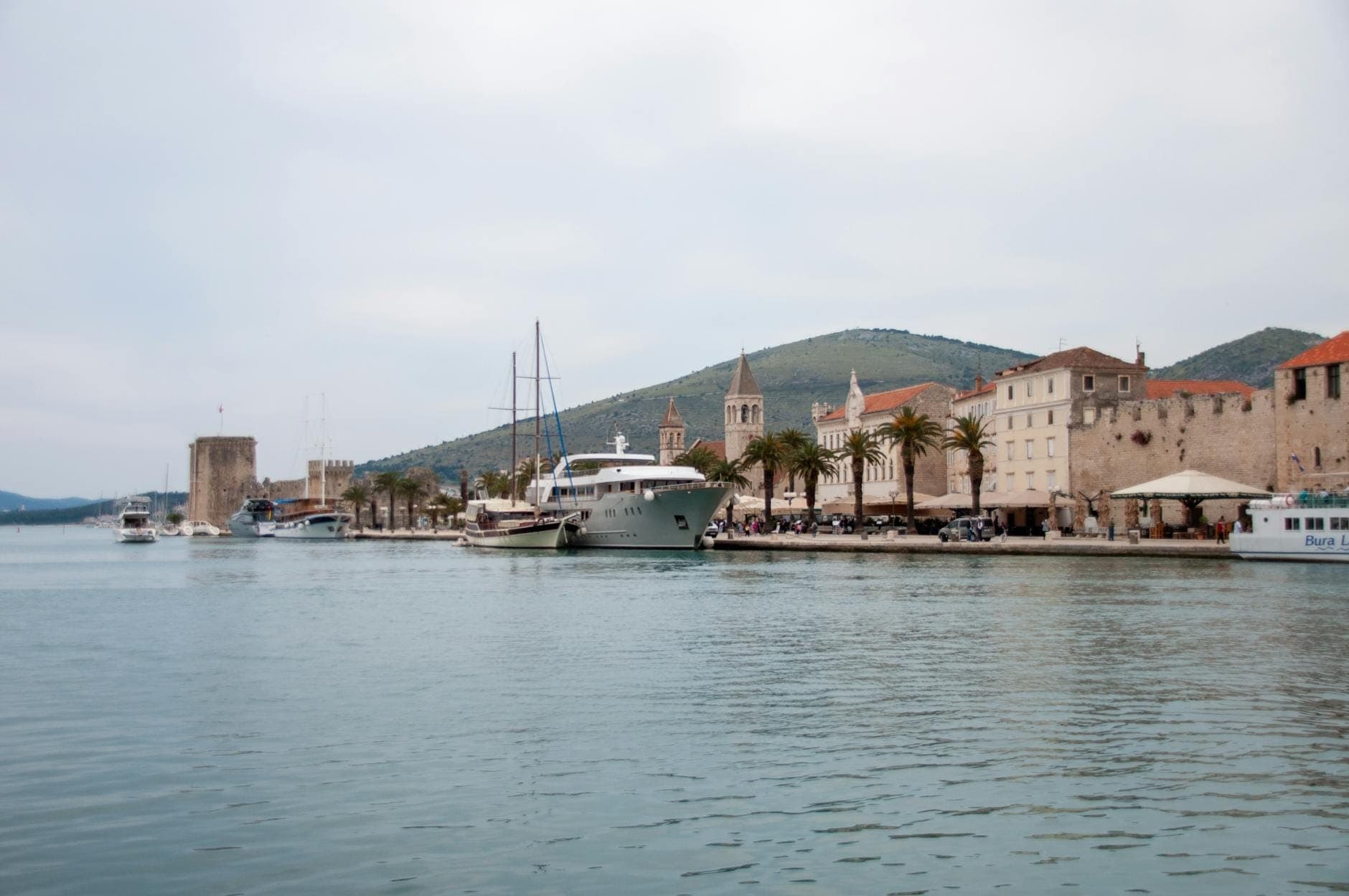 Vue du front de mer de Trogir avec ses bâtiments historiques en pierre, ses palmiers et ses bateaux amarrés dans le port par temps couvert.