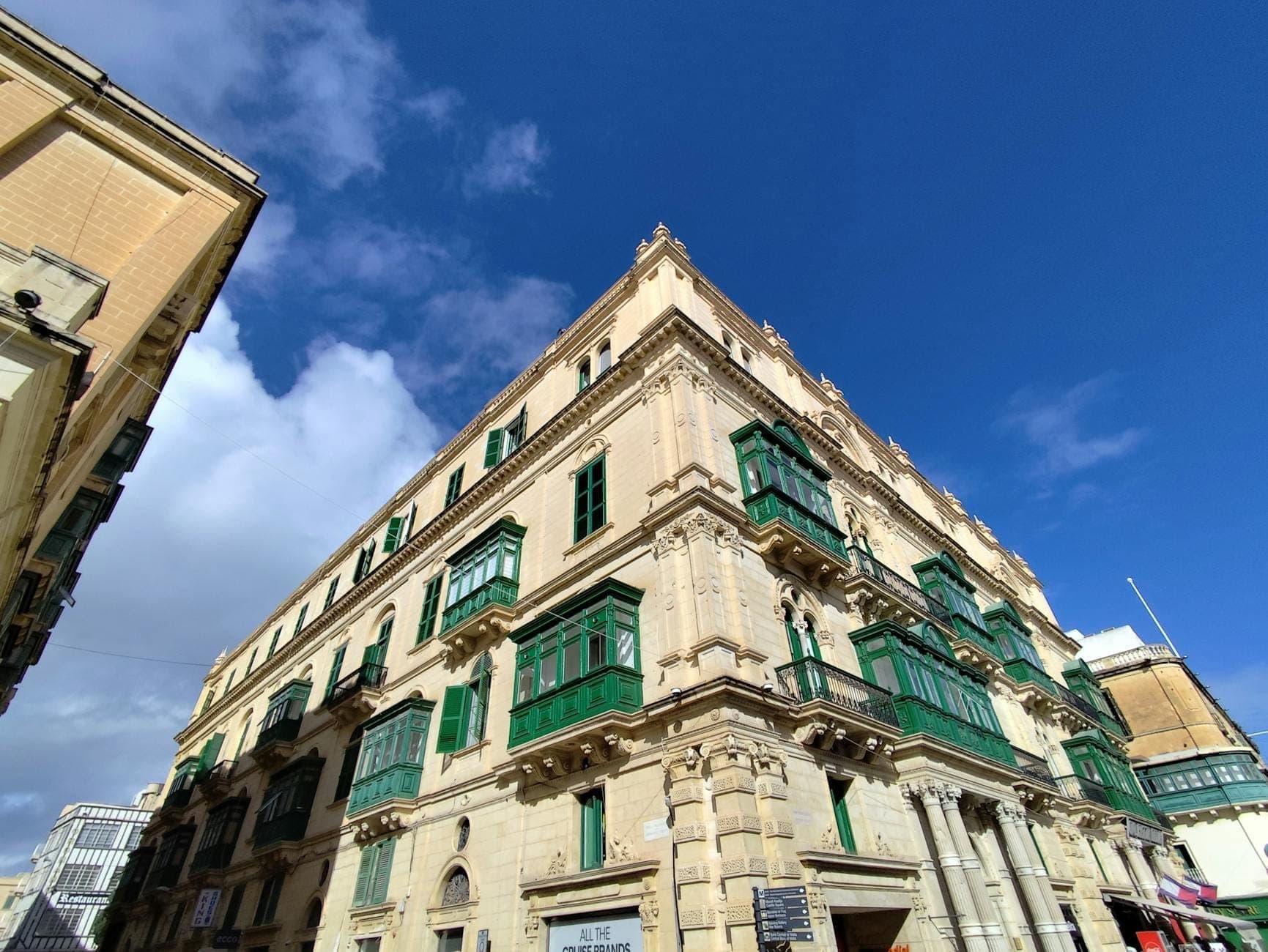 Large stone building with traditional Maltese green balconies under a blue sky on a city street corner in Valletta.
