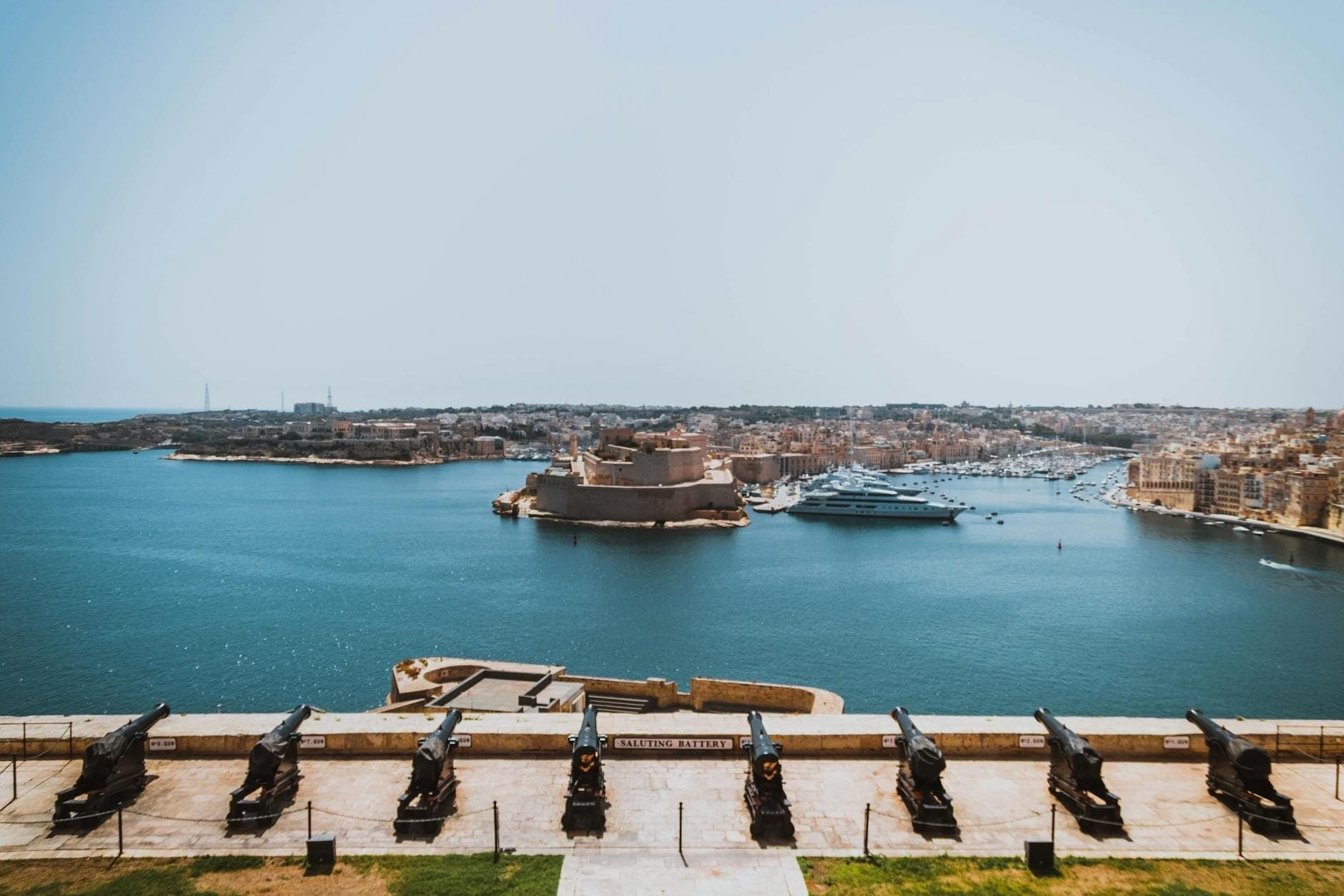 Panoramablick auf Vallettas Grand Harbour mit historischen Kanonen im Vordergrund und dem imposanten sternförmigen Fort St. Angelo auf der gegenüberliegenden Seite unter strahlend blauem Himmel.
