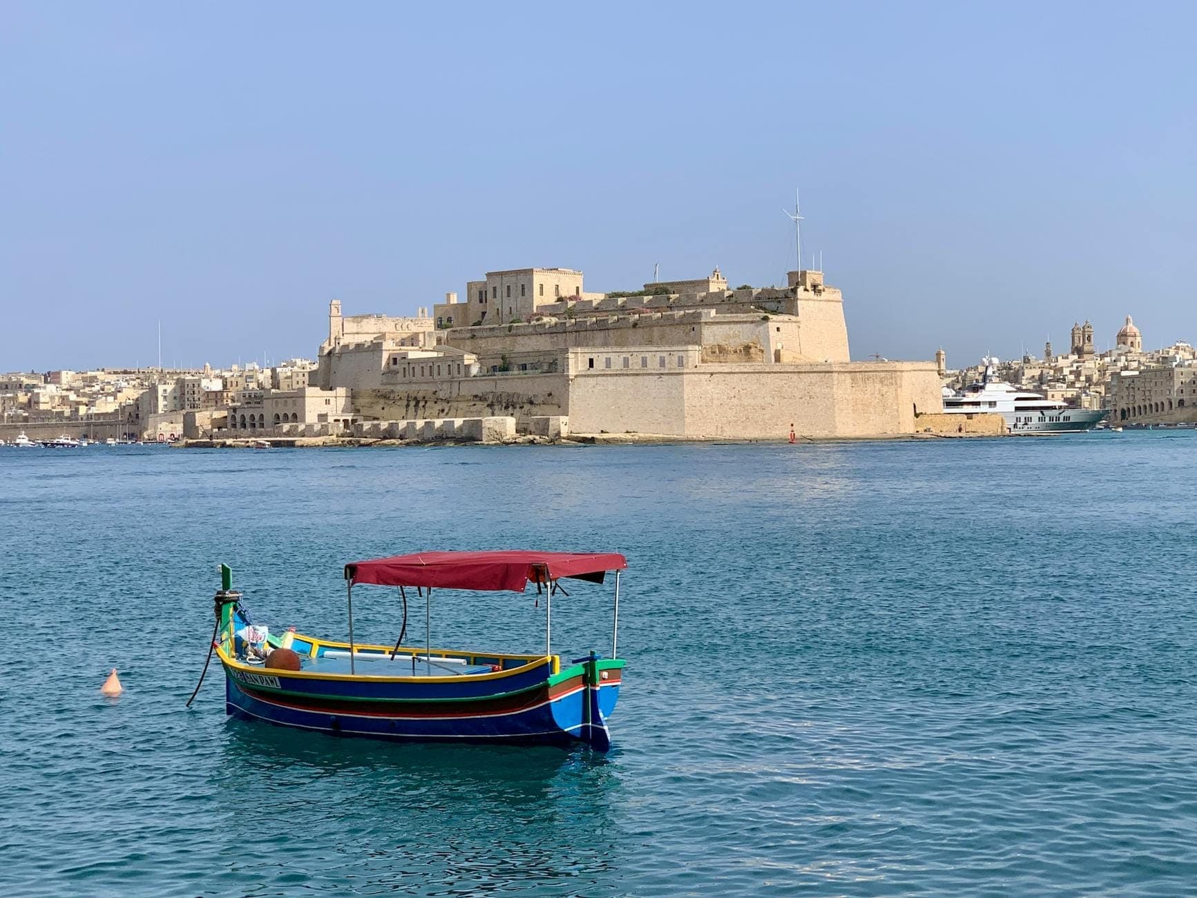 Traditional Maltese boat with red canopy floating on blue Grand Harbour water, with historic stone fortifications of Valletta in the background.