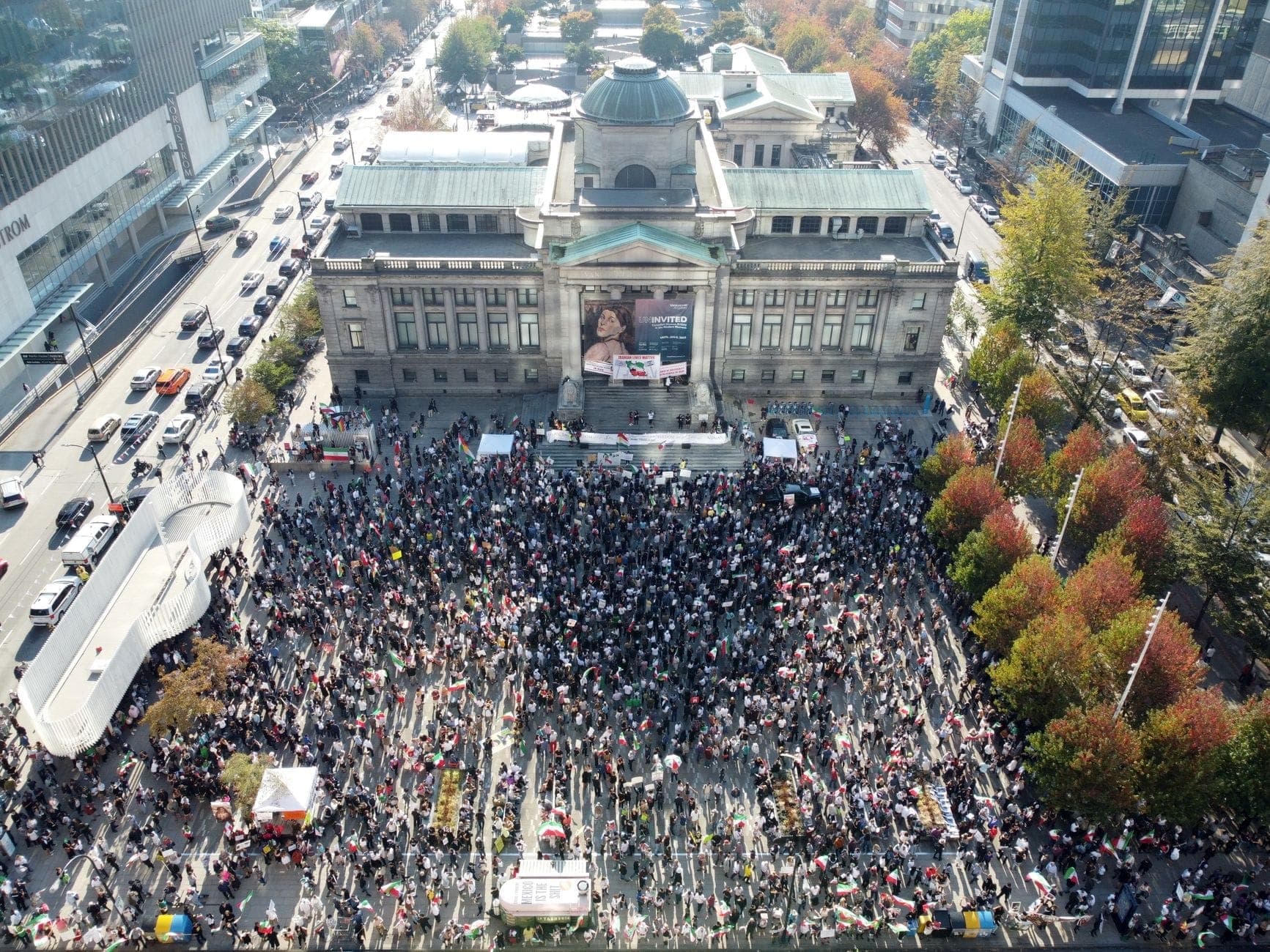 มุมมองทางอากาศของ Vancouver Art Gallery พร้อมฝูงชนจำนวนมากที่รวมตัวกันบนลานกลางเมือง Vancouver ล้อมรอบด้วยถนนในเมืองและต้นไม้ในฤดูใบไม้ร่วง