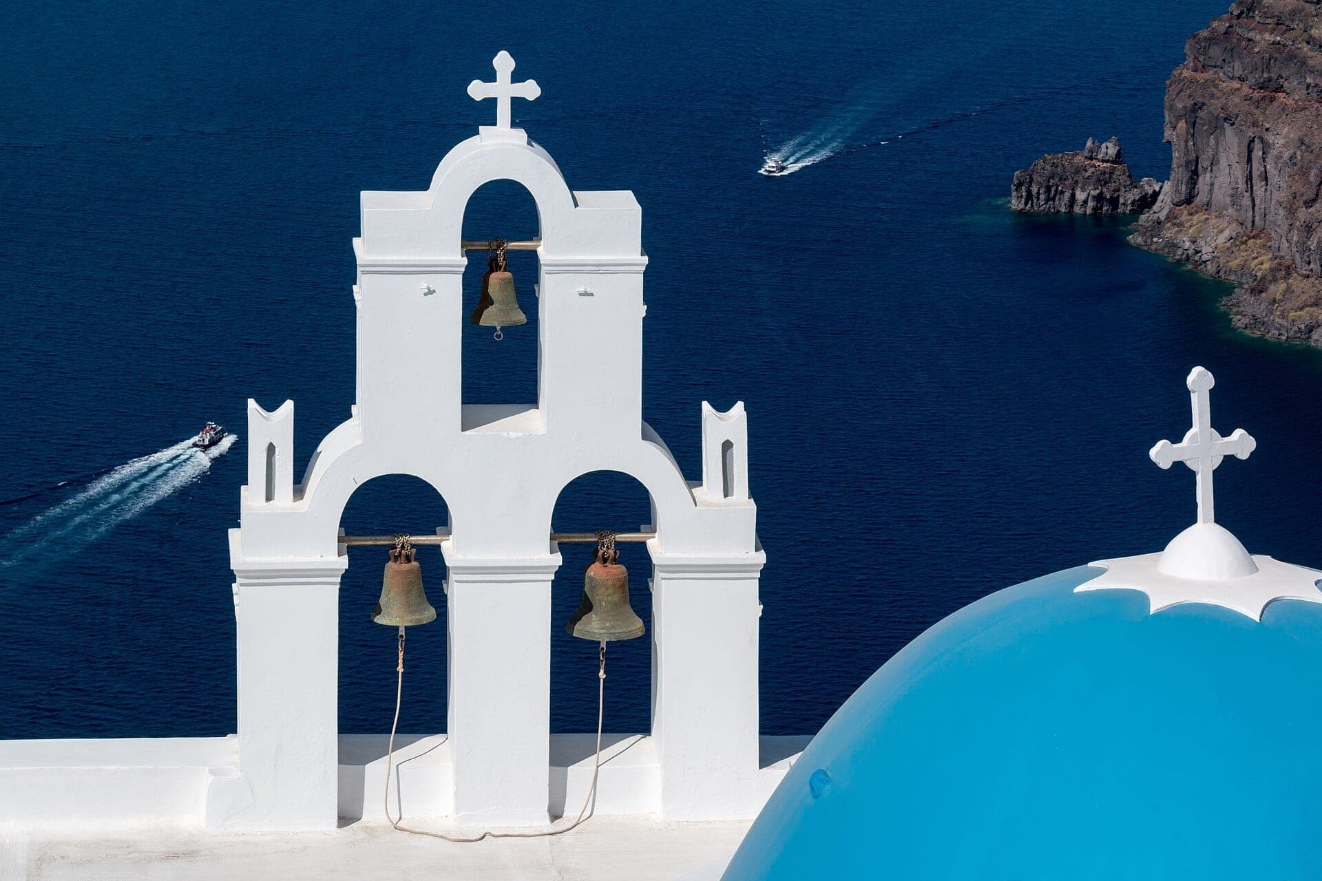 Die Drei Glocken der Kirche von Fira mit ihrem weißen Glockenturm, der blauen Kuppel und dem Ägäischen Meer im Hintergrund.