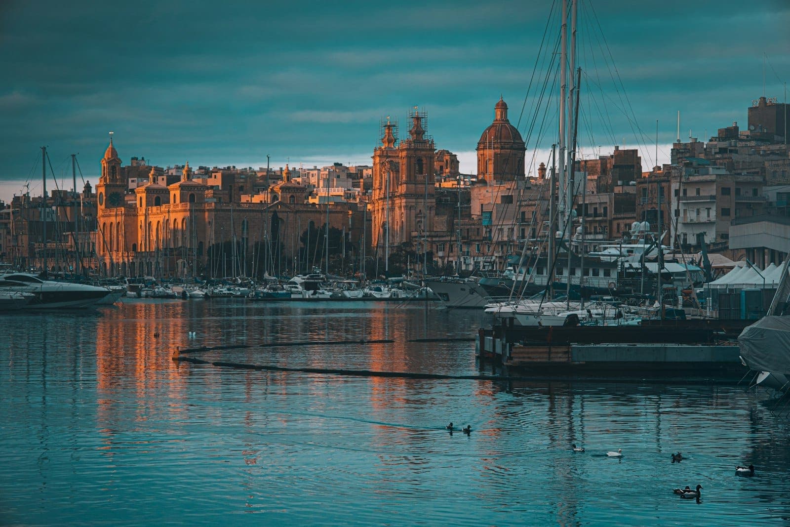 Goldenes Morgenlicht taucht das Ufer einer alten befestigten Stadt in Malta mit Kuppelkirchen, Booten und ruhigen Spiegelungen im Grand Harbour in warmes Licht.