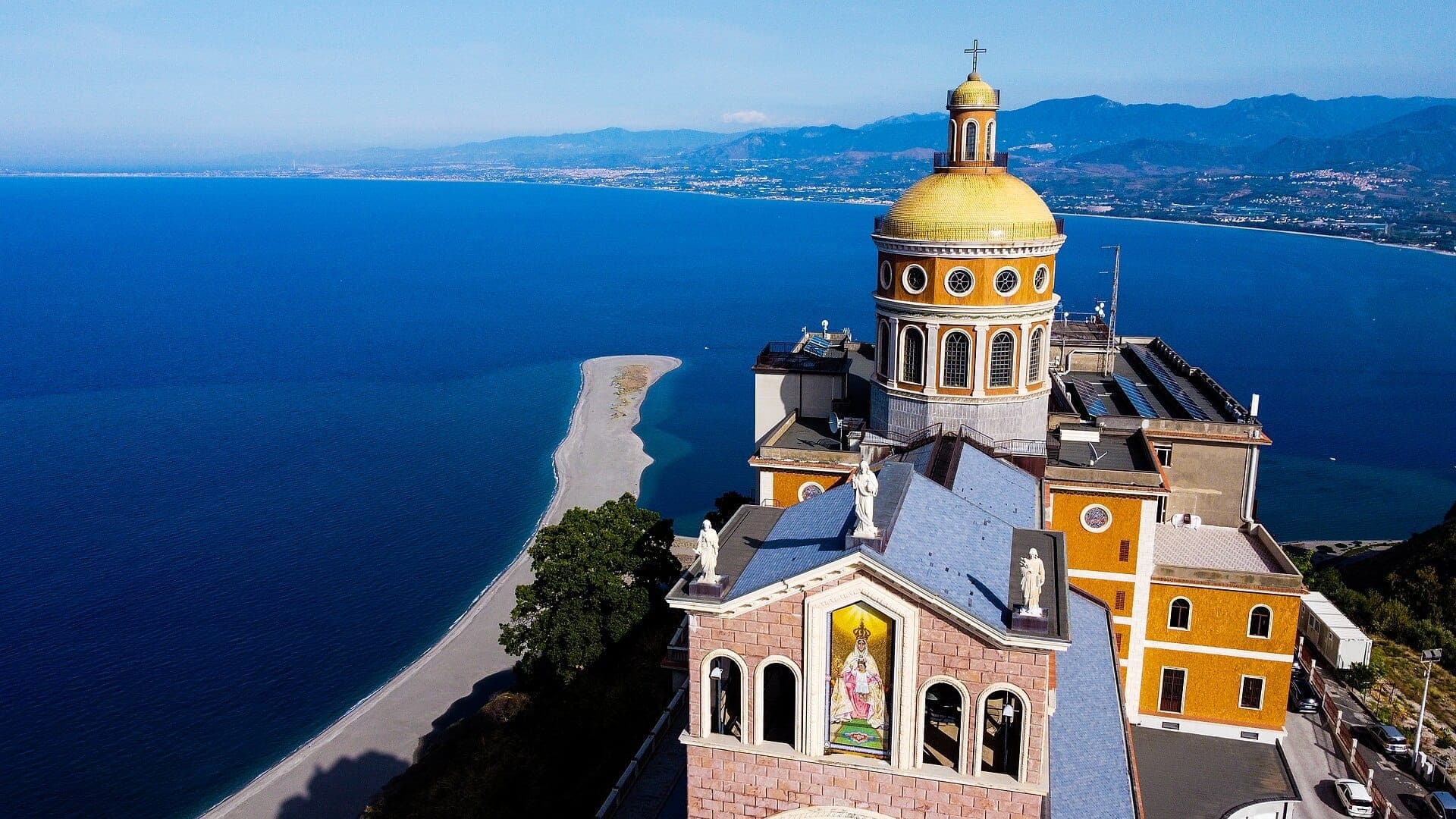 Pemandangan udara Sanctuary of Tindari dengan kubah emasnya, menghadap Laut Tyrrhenian biru cerah dan garis pantai Sisilia yang dramatis.