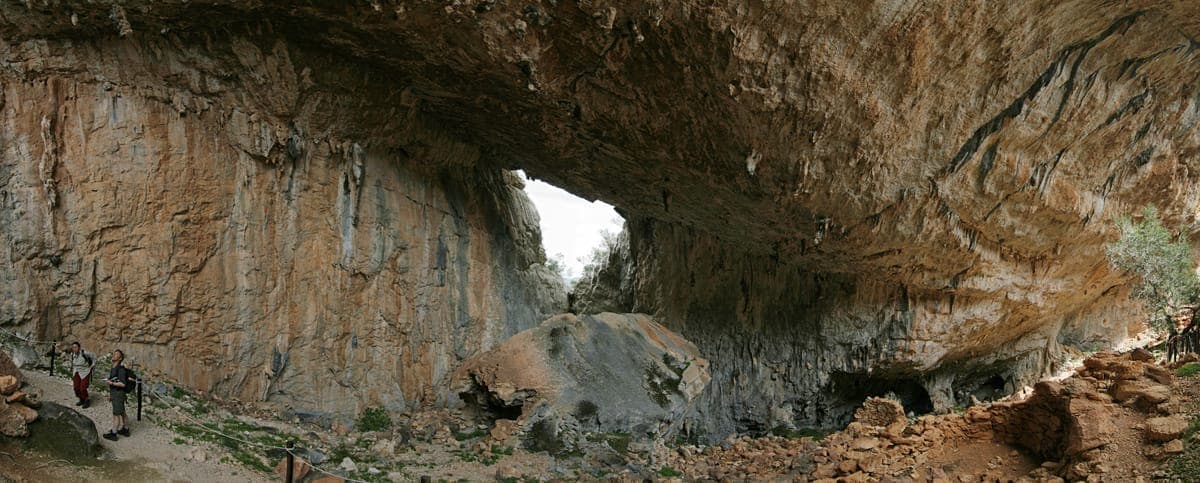 Gran abertura de cueva calcárea en el poblado nurágico de Tiscali, con dos senderistas caminando por un sendero rocoso bajo los acantilados en voladizo.
