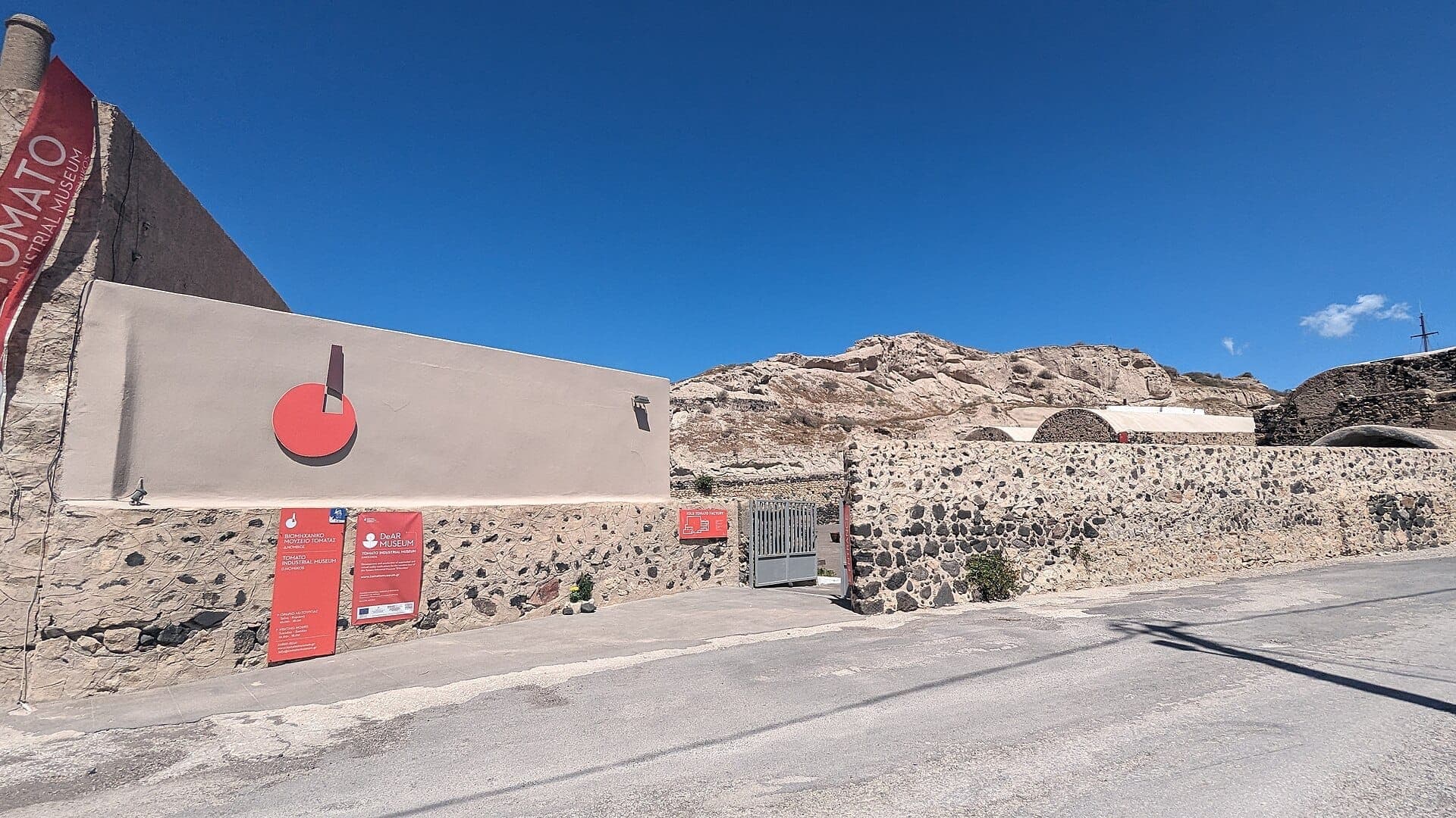 Haupteingang des Tomato Industrial Museum D. Nomikos in Santorini, mit Steinmauern, roter Beschilderung und felsigen Hügeln unter einem klaren blauen Himmel.