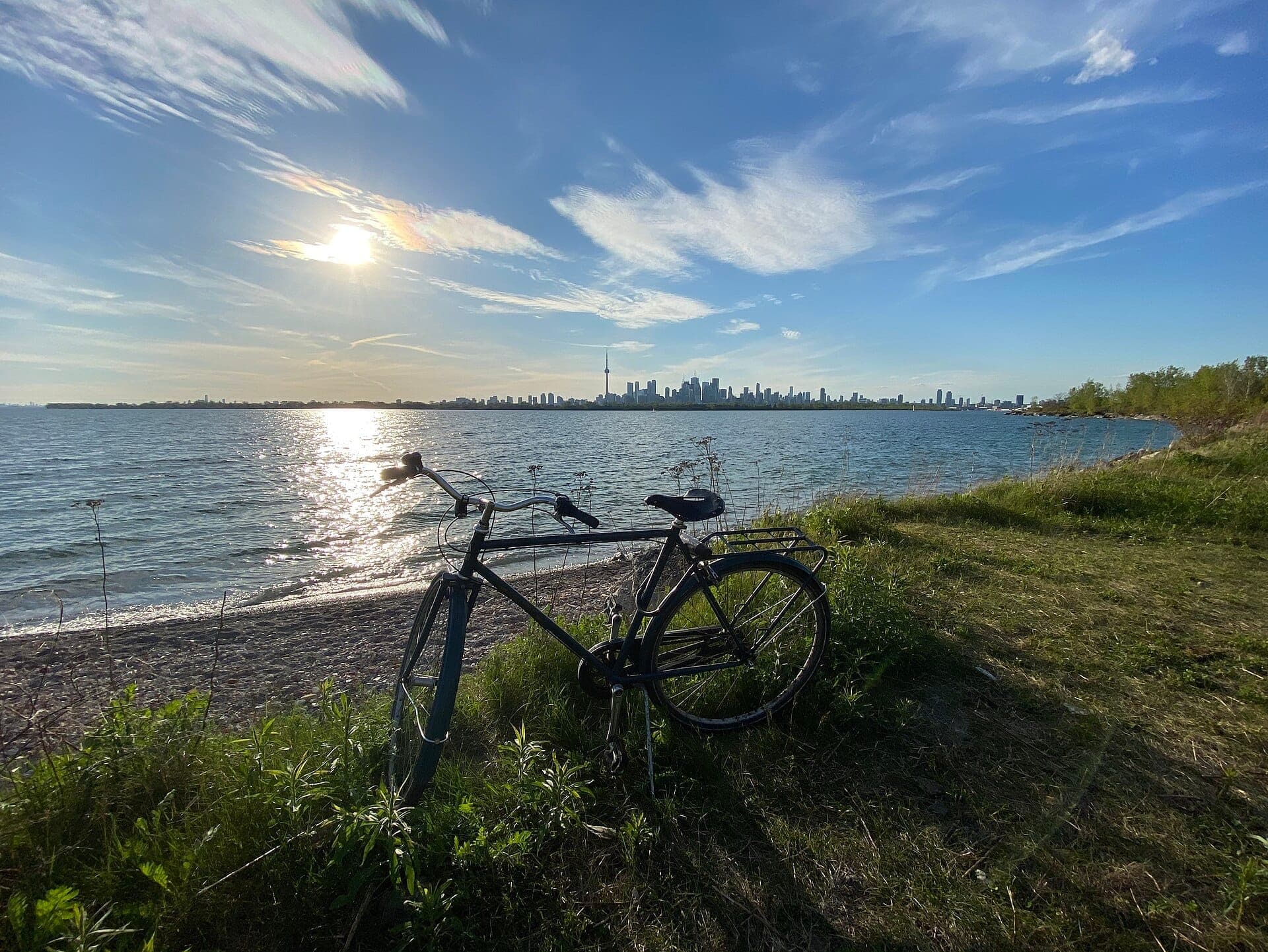 Bicicletta appoggiata sulla riva erbosa del Tommy Thompson Park con lo skyline di Toronto e il lago Ontario sotto un cielo parzialmente nuvoloso e soleggiato.