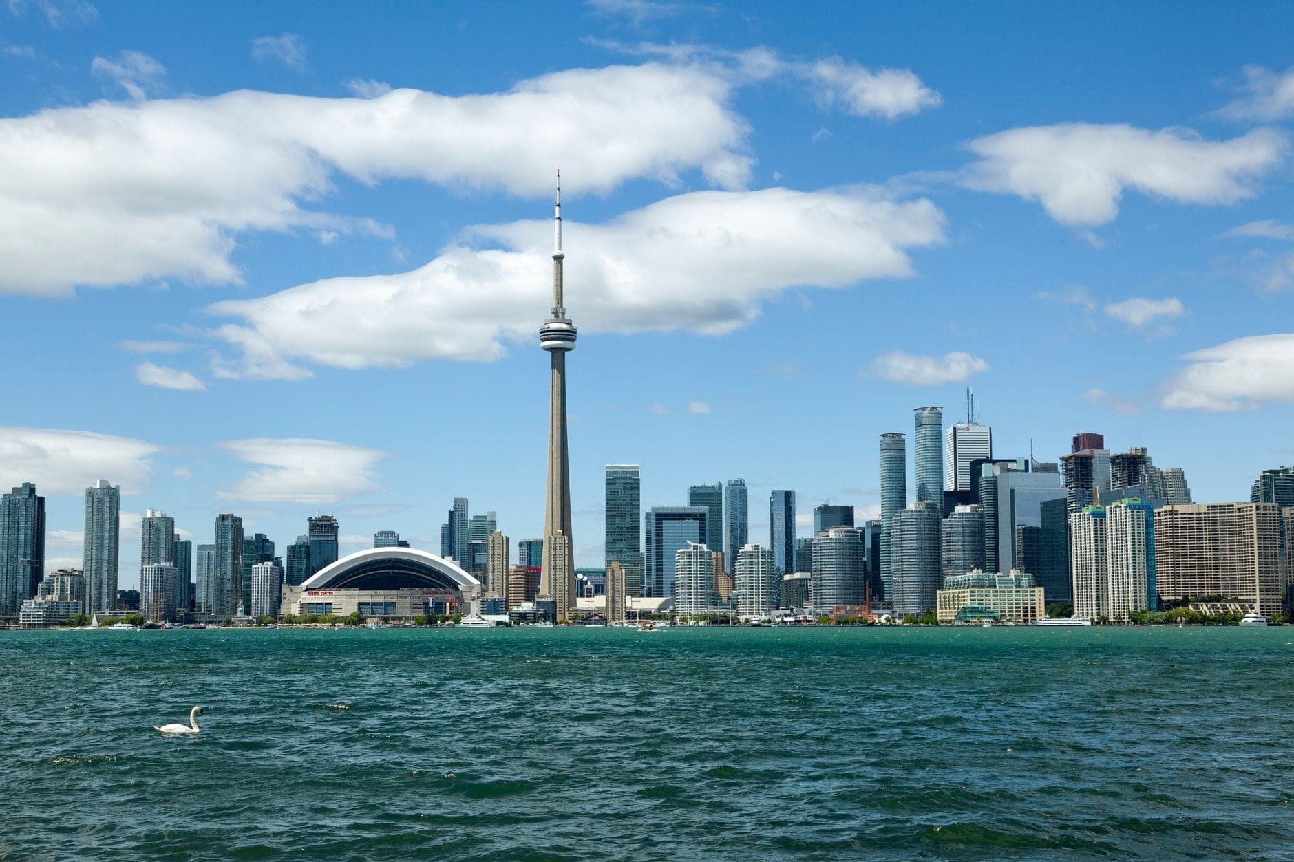 Cakrawala Toronto dengan CN Tower di hari yang cerah, dilihat dari seberang danau dengan langit biru dan awan putih bergelantungan di atas.