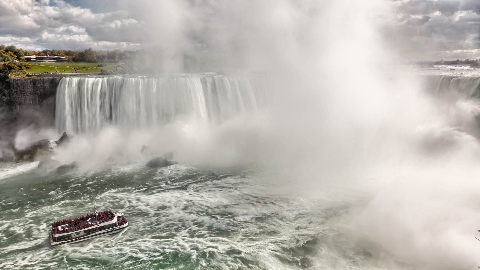 Vista amplia de las Cataratas del Niágara con un barco turístico en primer plano y niebla que se eleva de las poderosas cataratas.