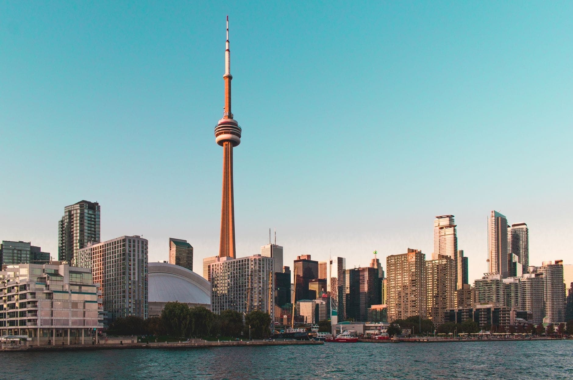 Cakrawala Toronto saat matahari terbenam dengan CN Tower, Rogers Centre, dan deretan gedung tinggi modern, cahaya hangat dengan Danau Ontario di latar depan.