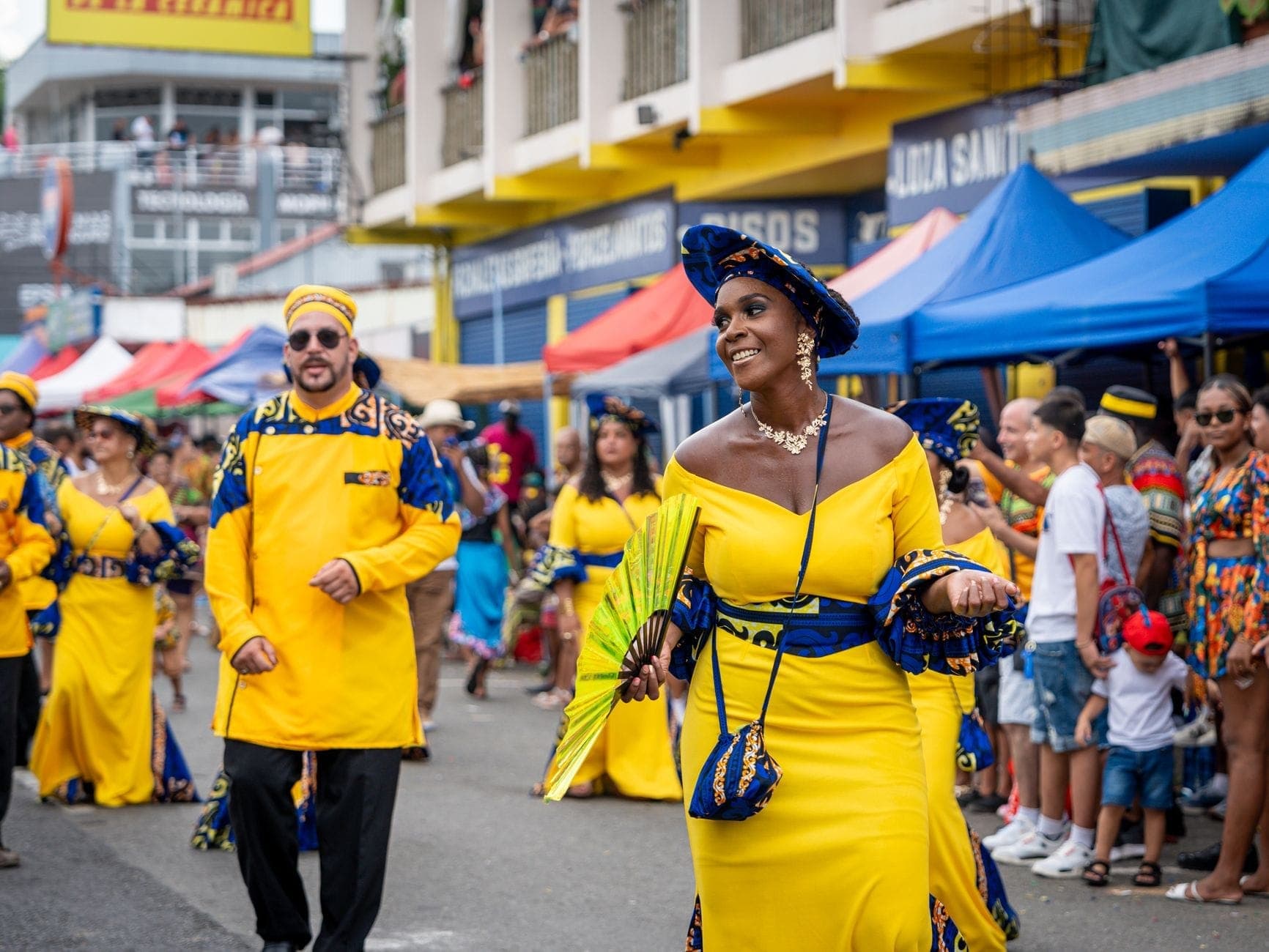 Peserta parade berpakaian kostum kuning cerah menari dan tersenyum sambil berjalan di sepanjang jalan kota yang dipenuhi tenda warna-warni dan penonton di festival yang meriah.