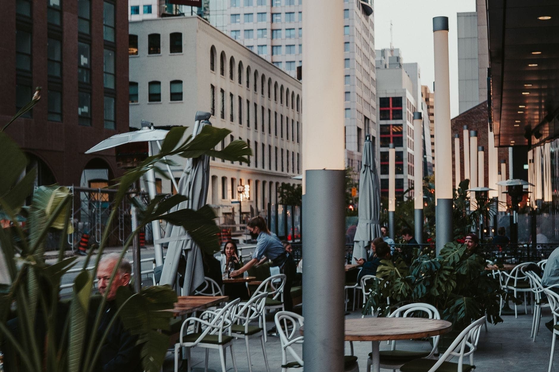 Patio di strada nel centro di Toronto con clienti ai tavoli circondati da edifici moderni e luci della città nel tardo pomeriggio.