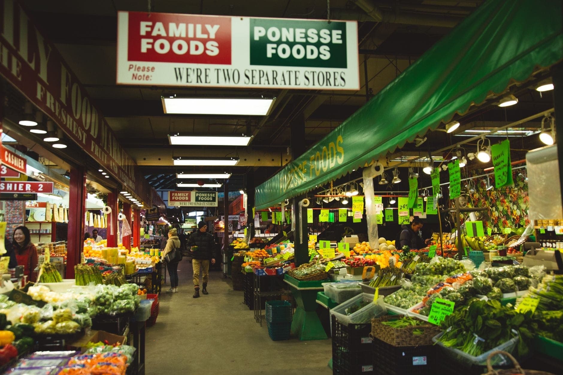 Mercato alimentare al coperto con bancarelle di prodotti freschi, verdure e frutta, acquirenti in giro e insegne in alto in un corridoio affollato.
