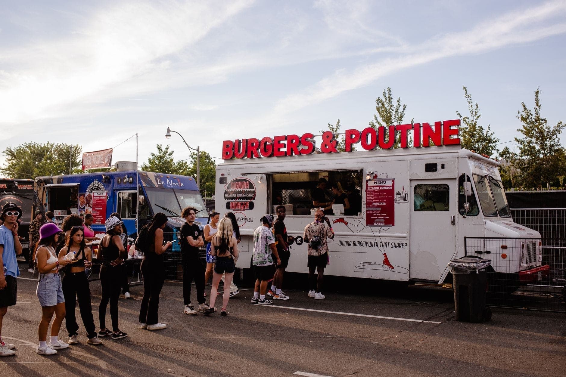 Orang-orang mengantri di food truck Toronto, termasuk sebuah truk putih dengan papan bertuliskan 'Burgers & Poutine' berwarna merah cerah, di bawah langit biru.