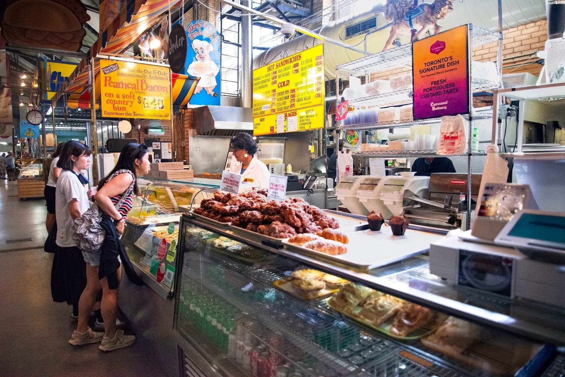 Brightly lit indoor food market with shoppers at a deli counter, glass display cases of food, and colorful signs overhead.