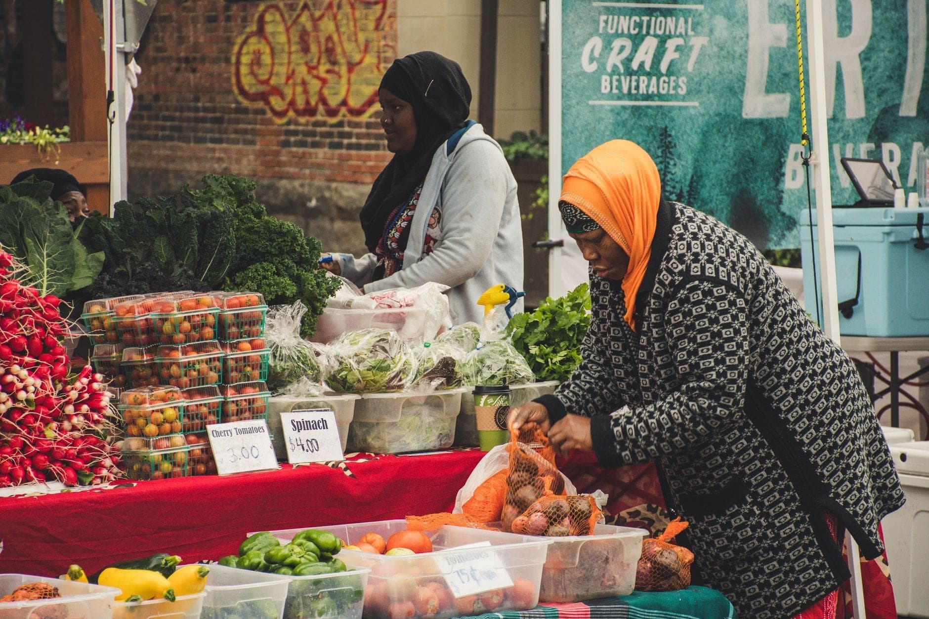 Two women selling fresh produce including tomatoes, greens, and peppers at a community market stall with handwritten signs.