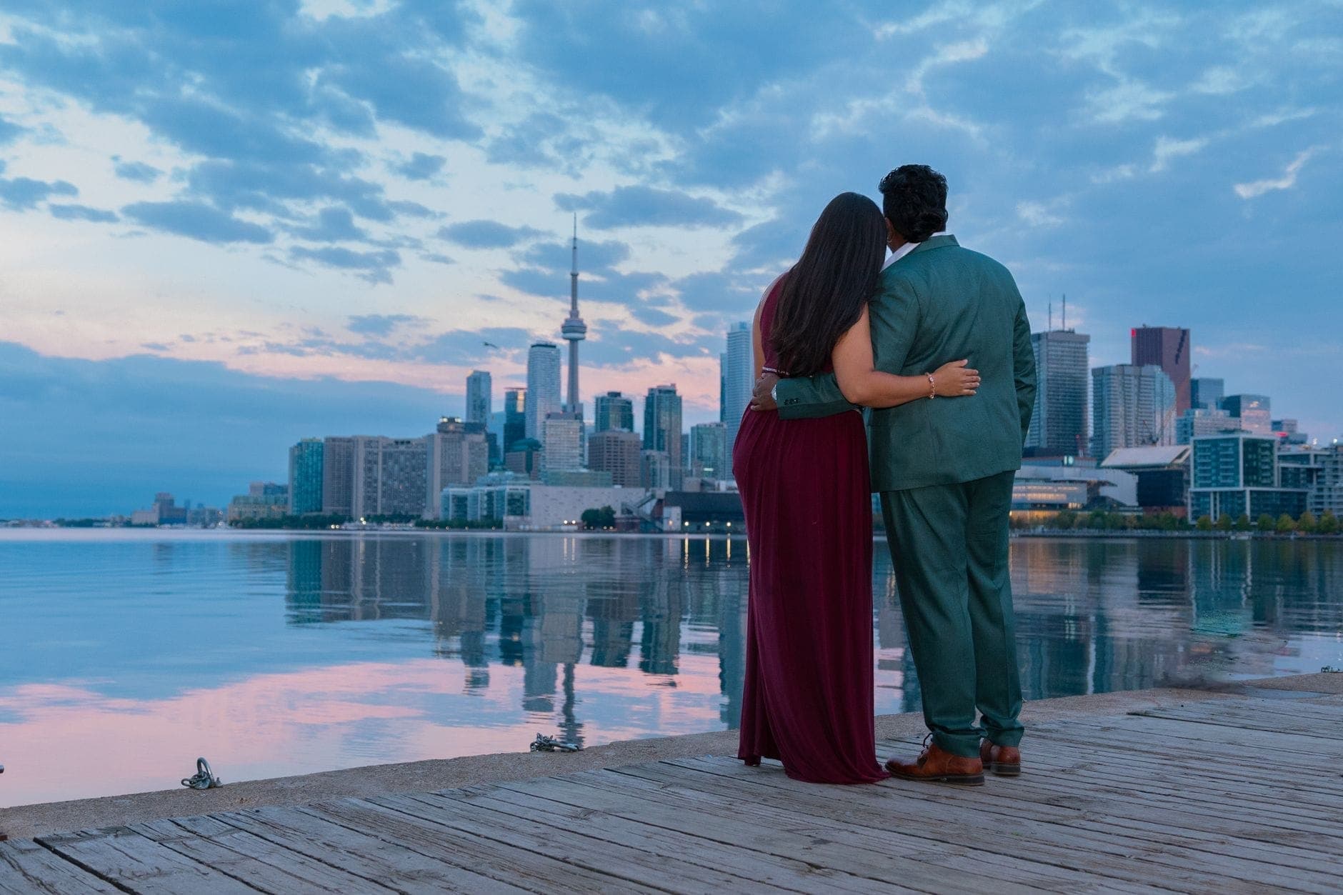 Pasangan berpelukan di tepi air Toronto saat senja, dengan skyline dan CN Tower di latar belakang yang terpantul di permukaan air tenang di bawah langit biru yang dramatis.