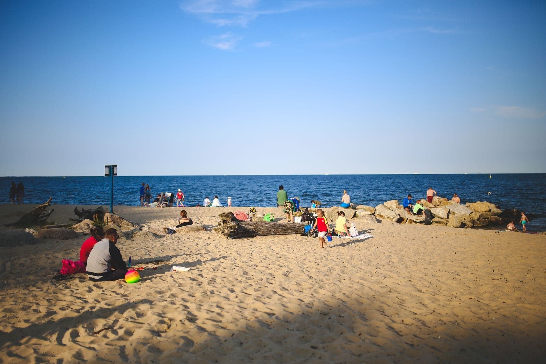 Persone che si rilassano e giocano su una spiaggia di sabbia vicino a un grande lago, con rocce e cielo azzurro limpido sullo sfondo.