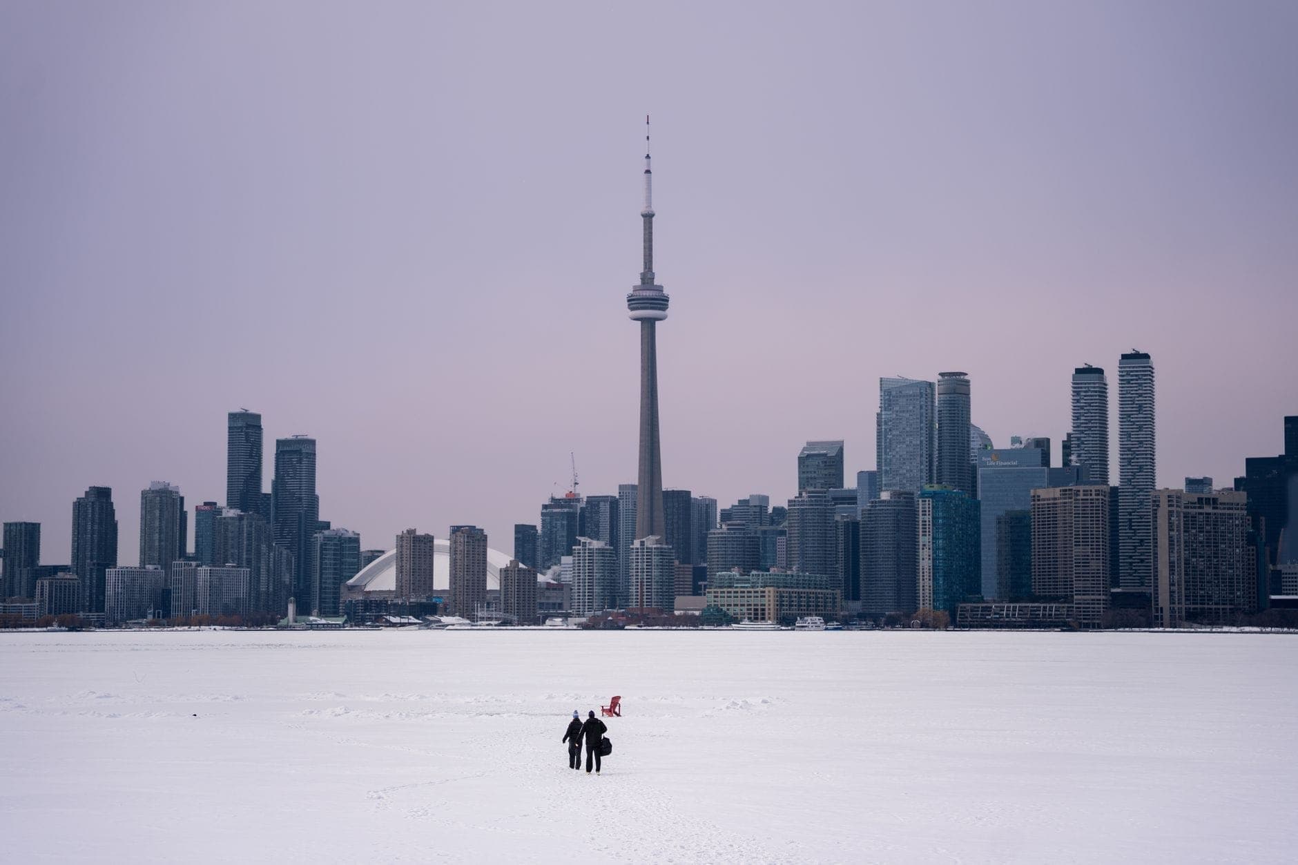 Dos personas caminando sobre un lago helado cubierto de nieve hacia el horizonte de Toronto y la CN Tower bajo un cielo invernal.