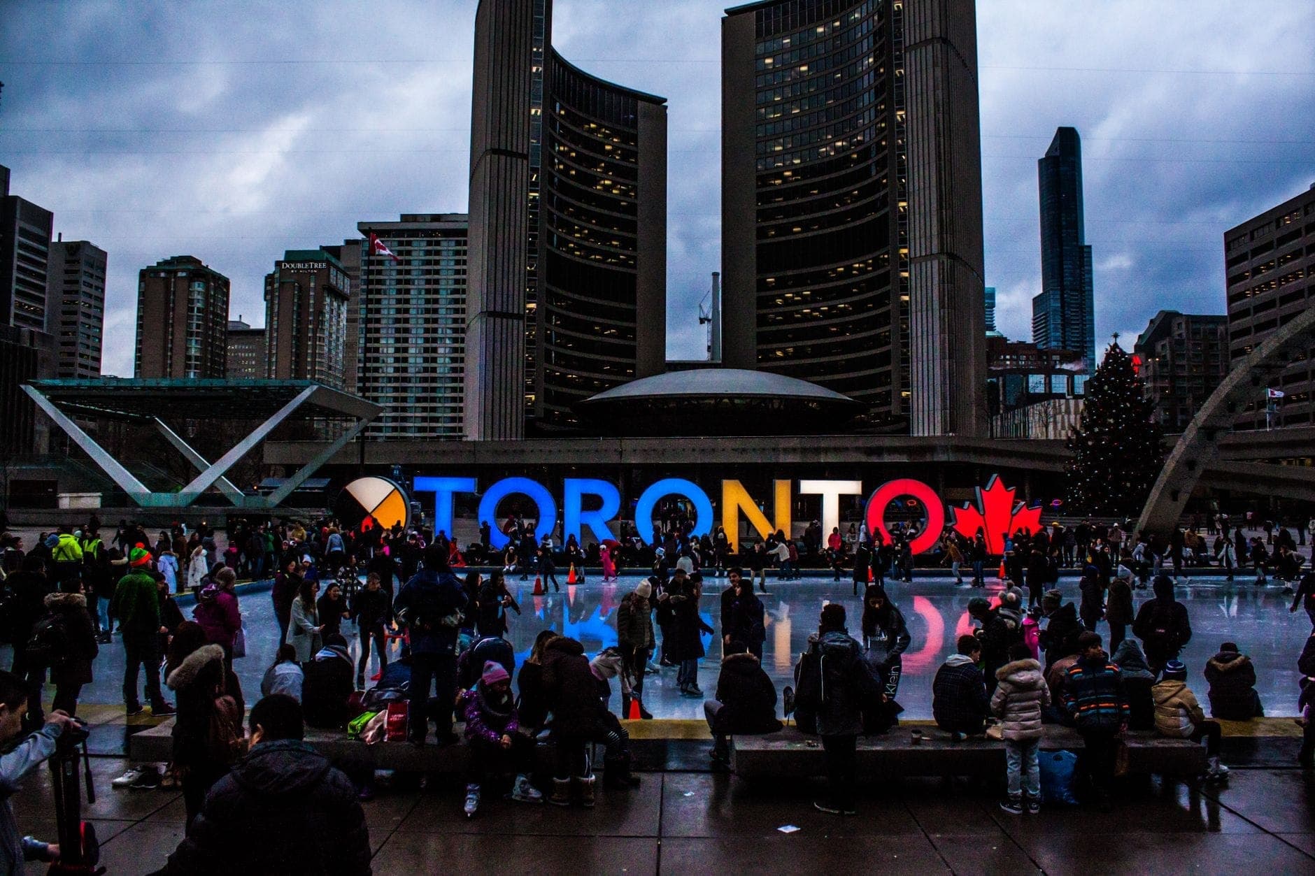 Crowd gathered at Nathan Phillips Square in front of the illuminated TORONTO sign with city hall buildings visible in the background.