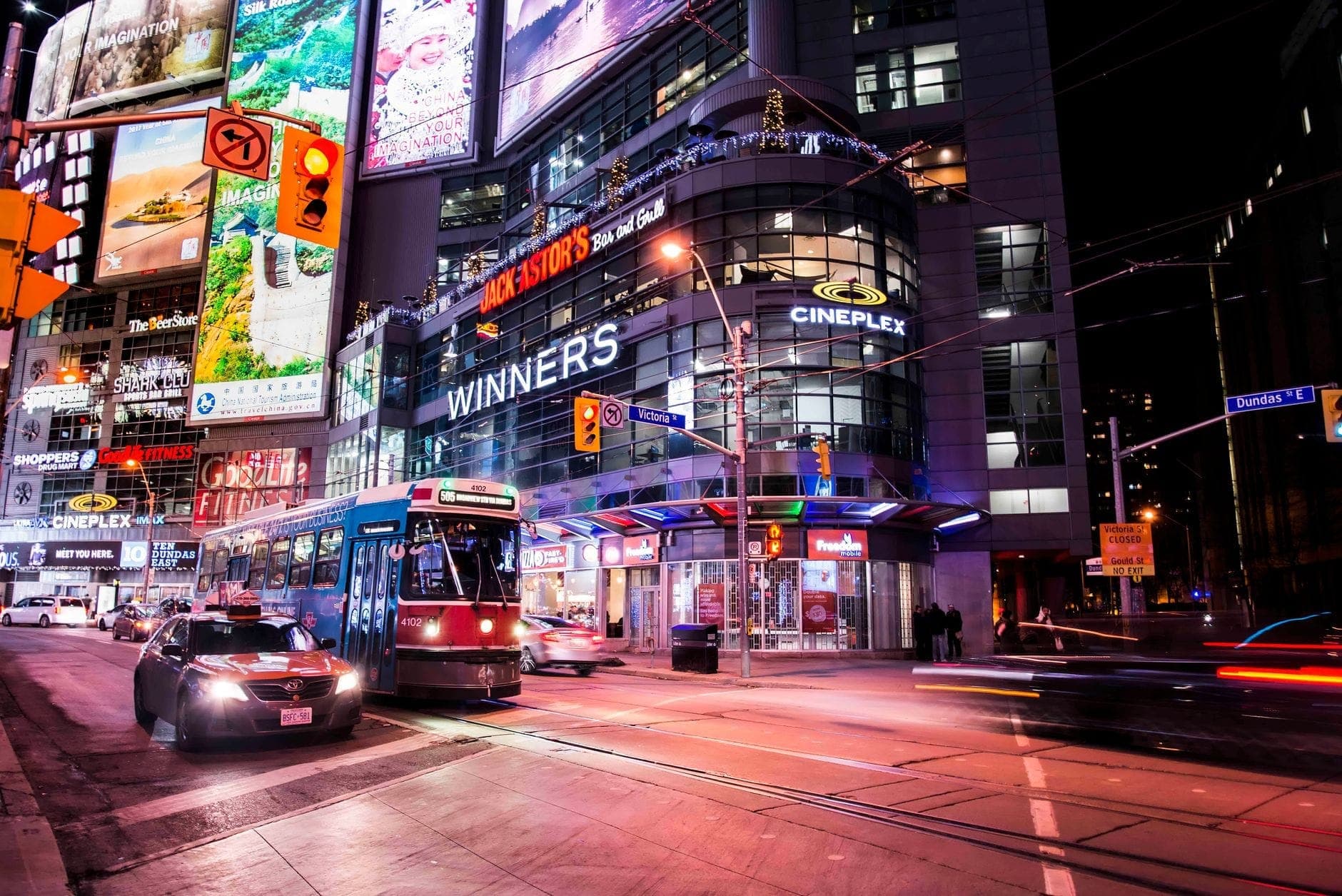 Streetcar passing by Cineplex and Winners at busy intersection in downtown Toronto at night with bright illuminated billboards and city bustle.