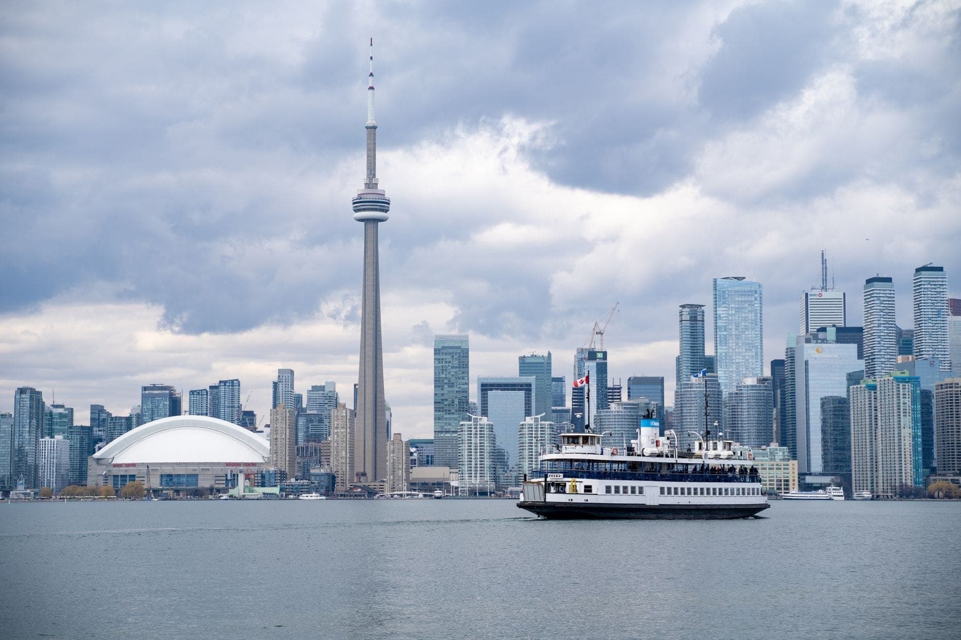 Toronto ferry crossing the water with the CN Tower and downtown skyline in the background under a cloudy sky.
