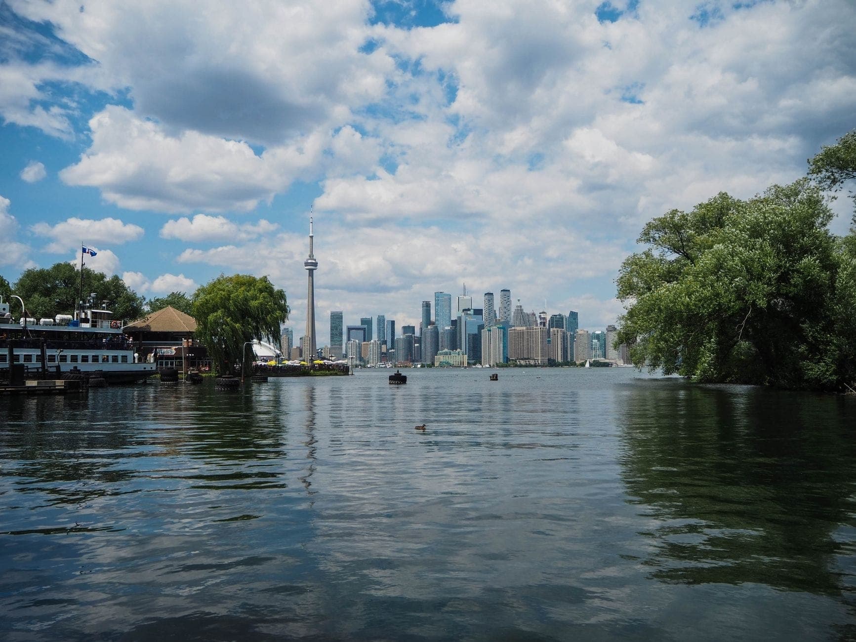 A view from the Toronto Islands showing trees, water, a ferry dock, and the downtown Toronto skyline with CN Tower on a partly cloudy day.