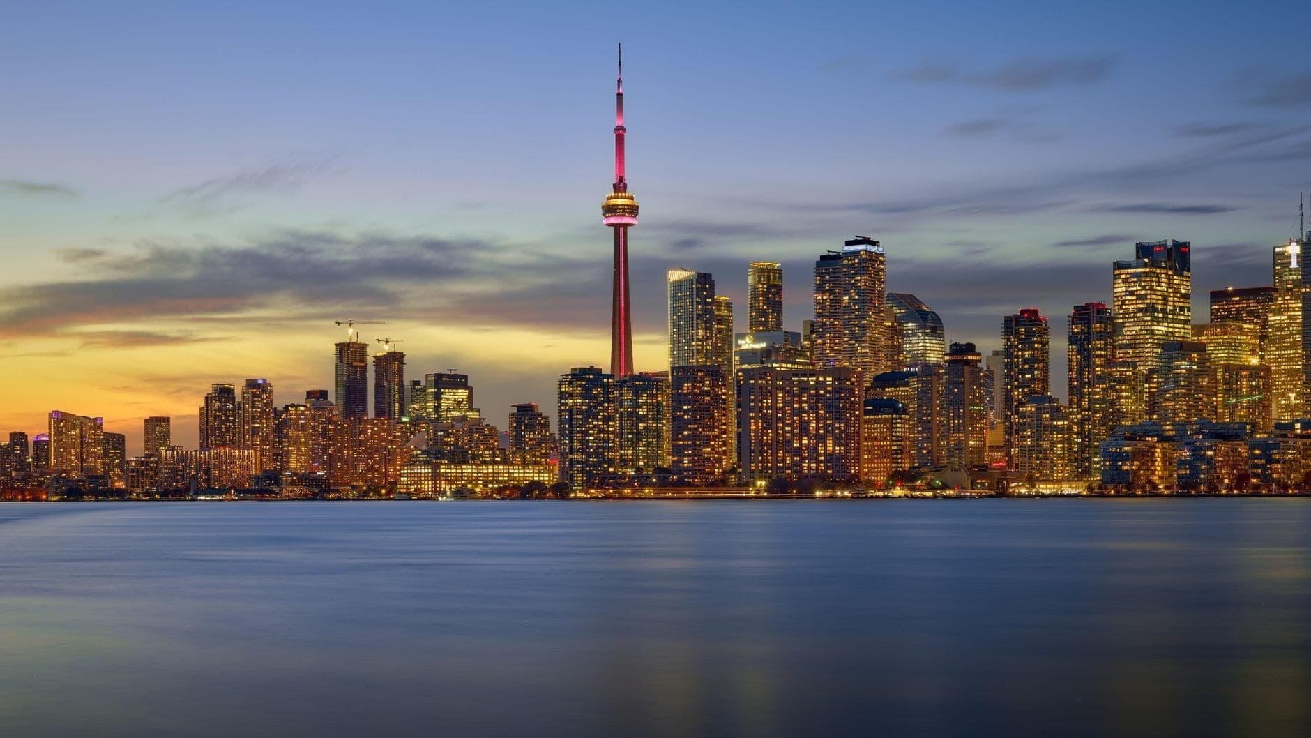 Pemandangan cakrawala Toronto saat senja dengan Menara CN dan cahaya kota yang memantul di Danau Ontario, memancarkan kesan mewah dan elegan.