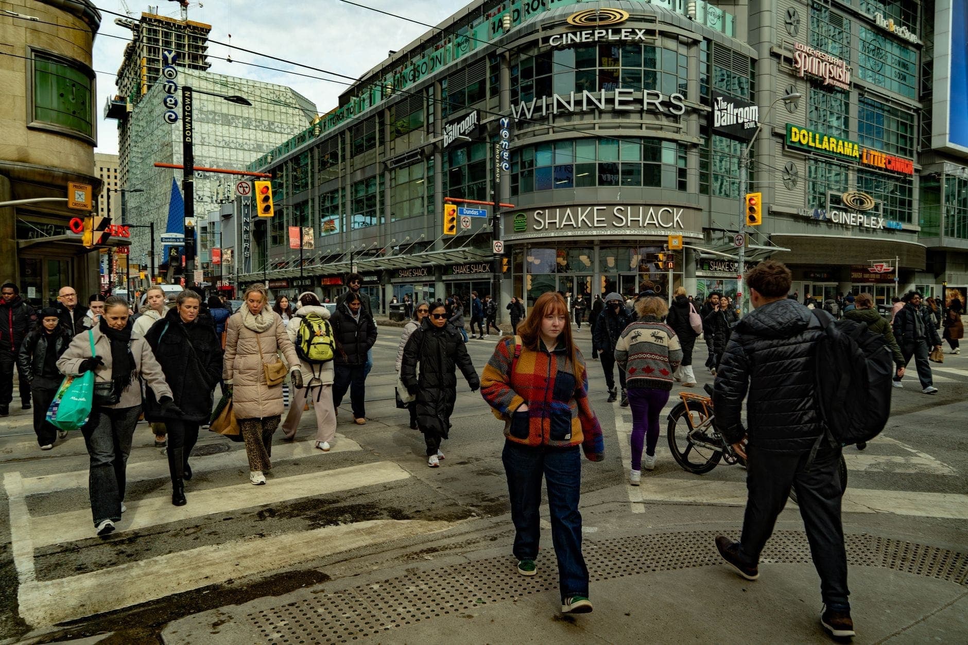Trafficato incrocio di Toronto con una folla eterogenea che attraversa davanti a negozi riconoscibili ed edifici urbani.