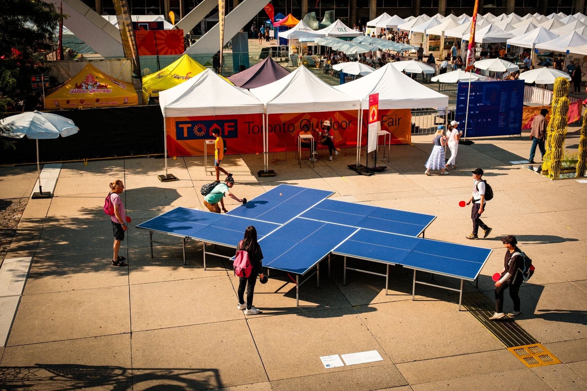 Persone che giocano a ping pong in una fiera di strada all'aperto a Toronto, fiancheggiata da tendoni del festival in una soleggiata giornata estiva.